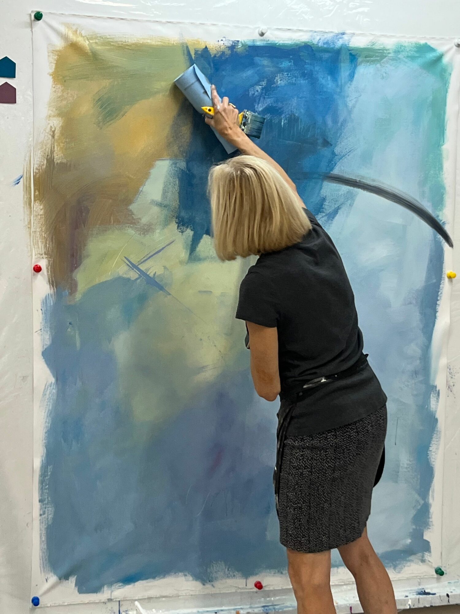 Child with blonde hair painting on a large canvas, wearing a black shirt and shorts, with colorful background.