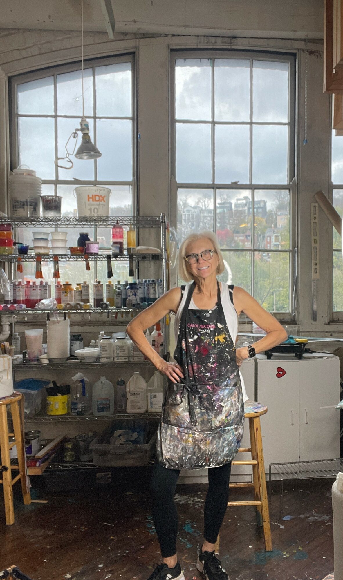 Woman with glasses wearing an apron stands in an art studio with large windows, shelves, and art supplies behind her.