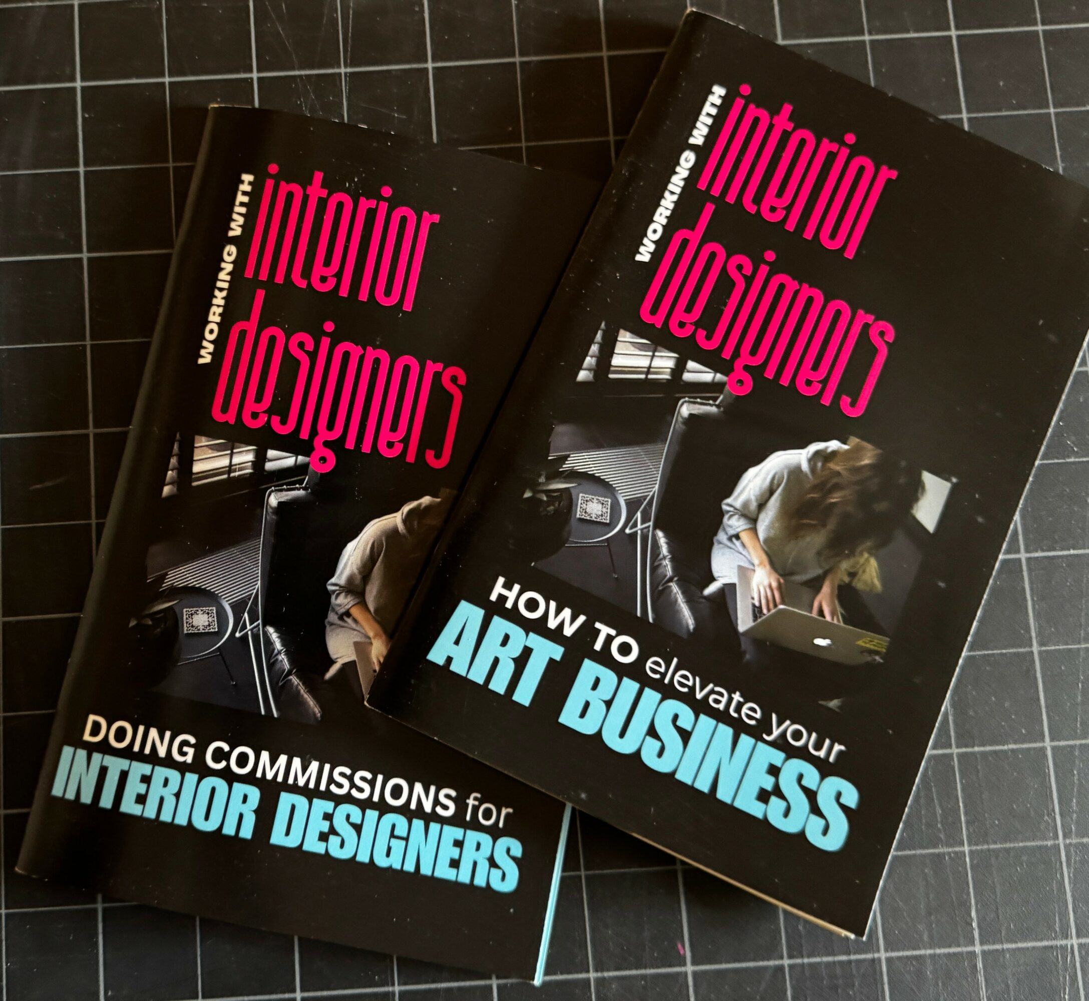 Two books titled 'Interior Designers' and 'How to elevate your Art Business' on a black tiled surface.