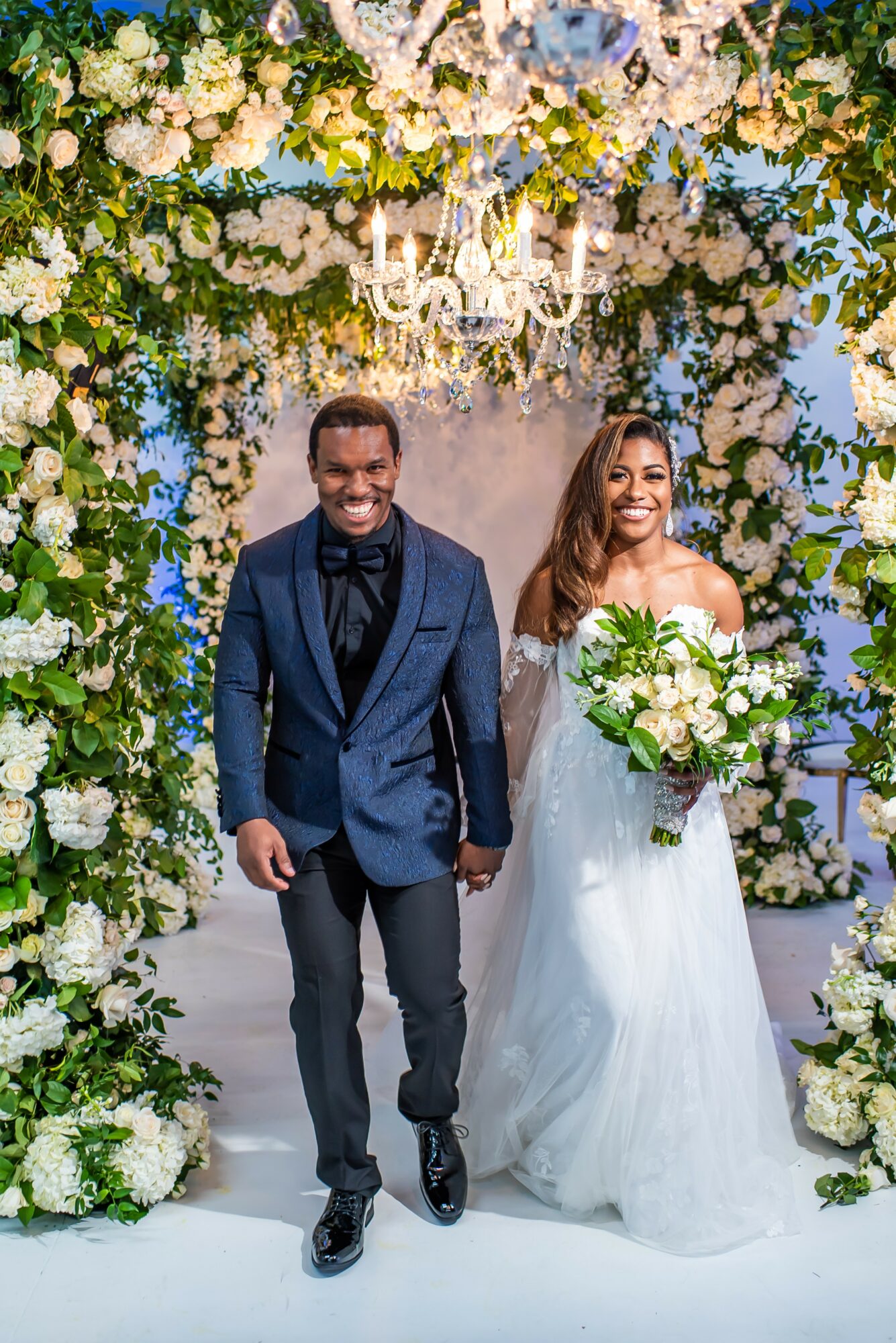 A smiling couple walks down an aisle decorated with white flowers and greenery, with a chandelier overhead, during a wedding.