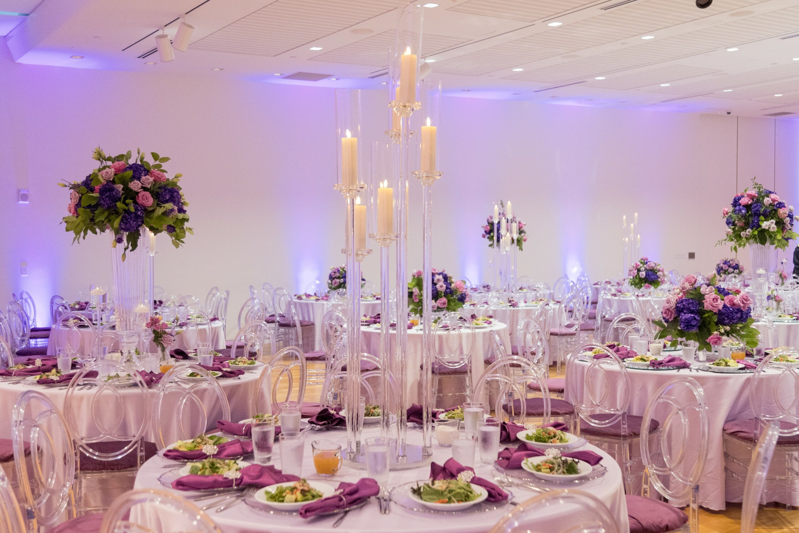 Elegant banquet hall with round tables, floral centerpieces, tall candles, and soft purple lighting, set for a formal event.