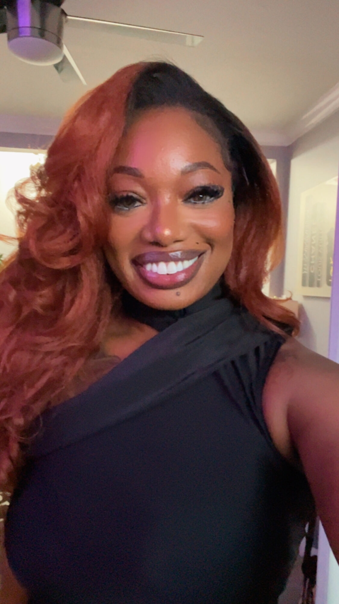 Woman with long, wavy reddish-brown hair smiling, wearing a black top, indoors with a ceiling light visible.