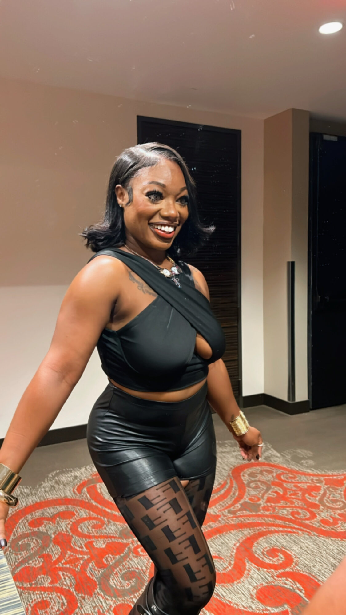 Woman smiling, wearing black top, leather shorts, patterned tights, in indoor setting with carpet and walls