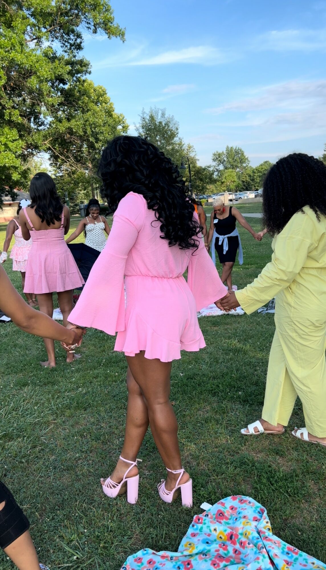 Group of women holding hands in a circle outdoors, with trees and a blue sky in the background.
