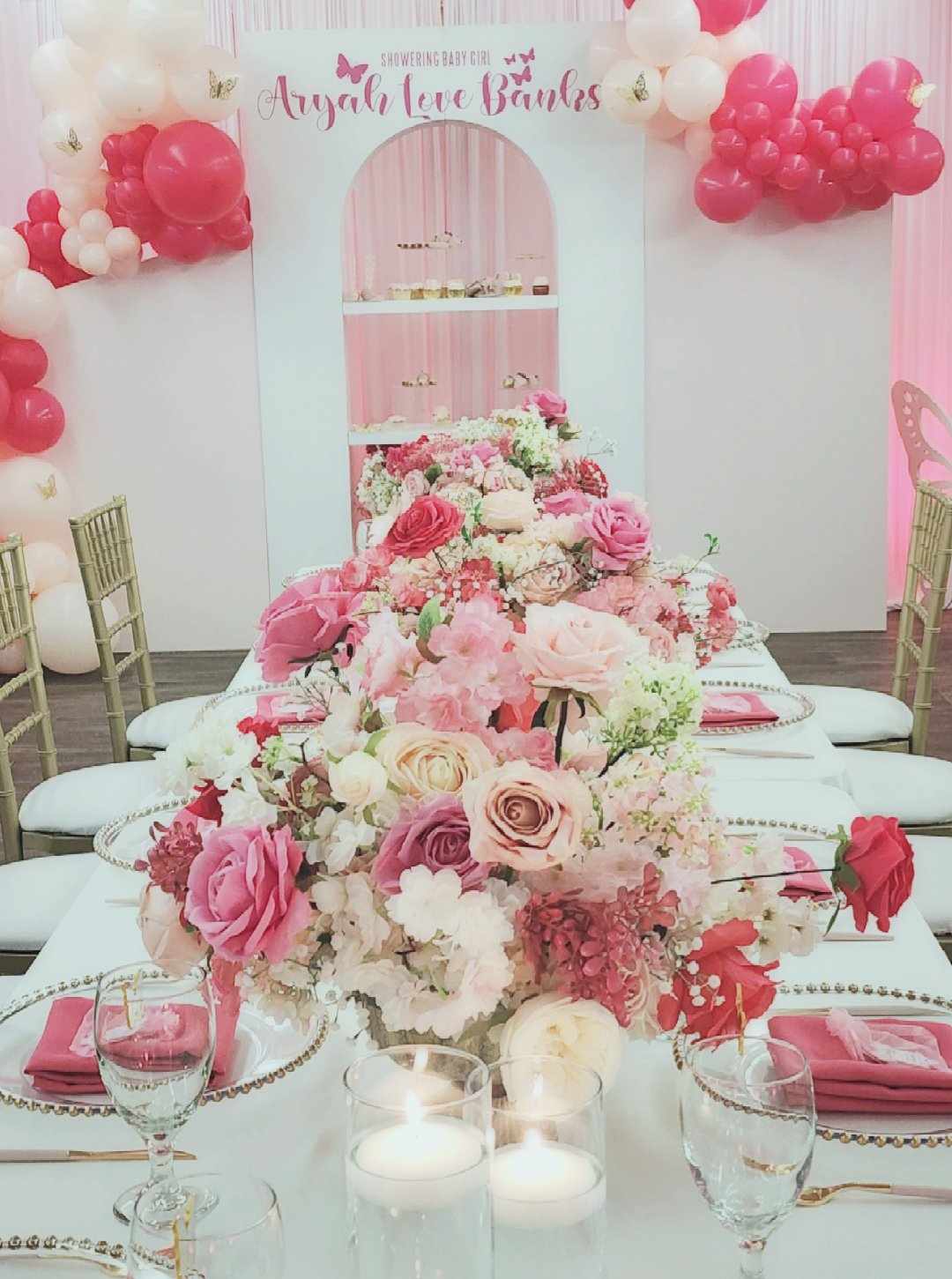 Decorated table with pink and white flowers, candles, and glassware in a festive setting, with pink and white balloons and a sign in the background.