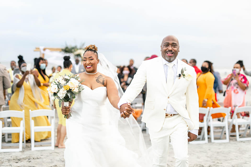Bride and groom holding hands, walking outdoors with guests in the background, bride holding a bouquet, smiling, wedding celebration