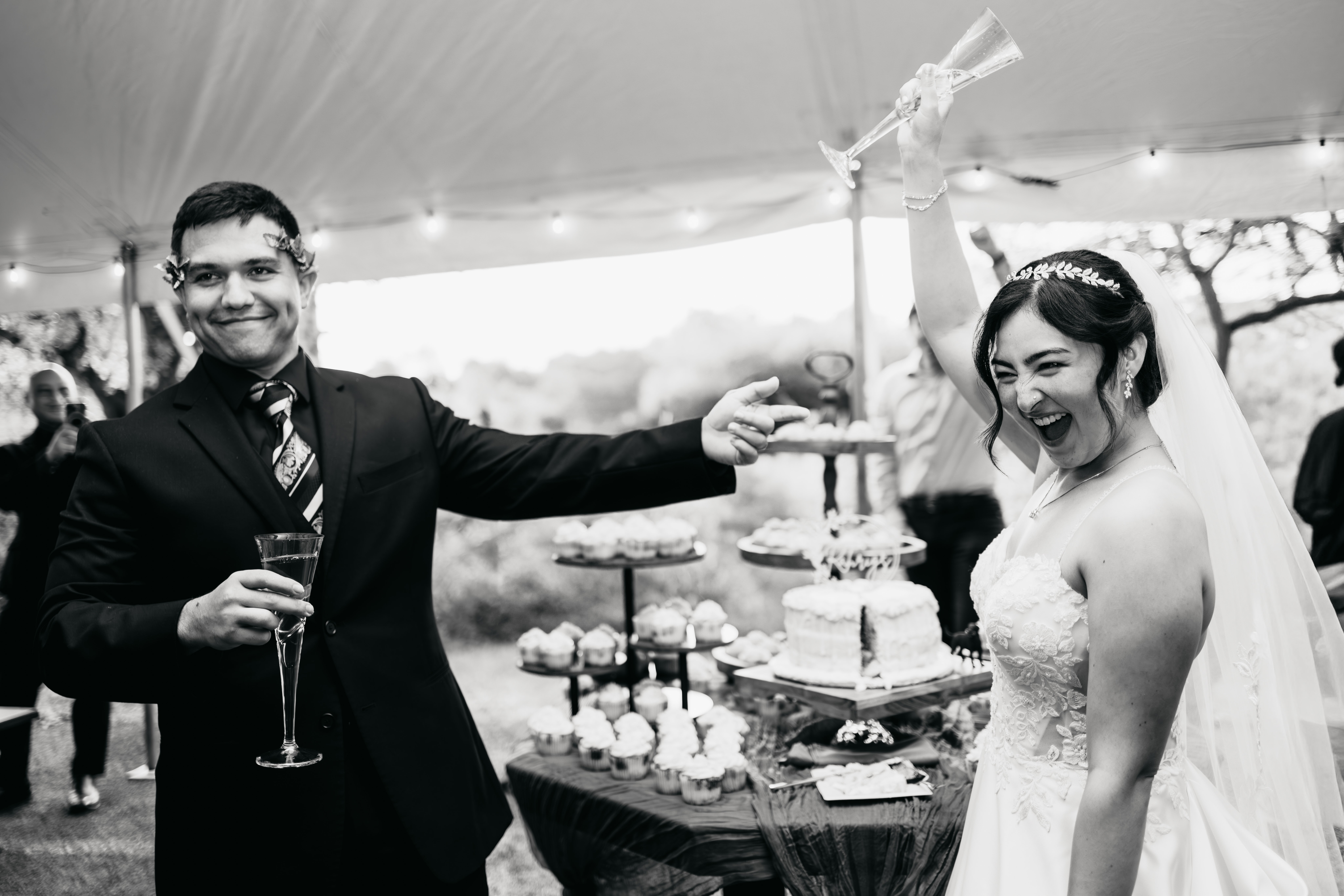 Man in suit and woman in wedding dress celebrate with raised hands and smiles at a wedding reception, holding drinks.