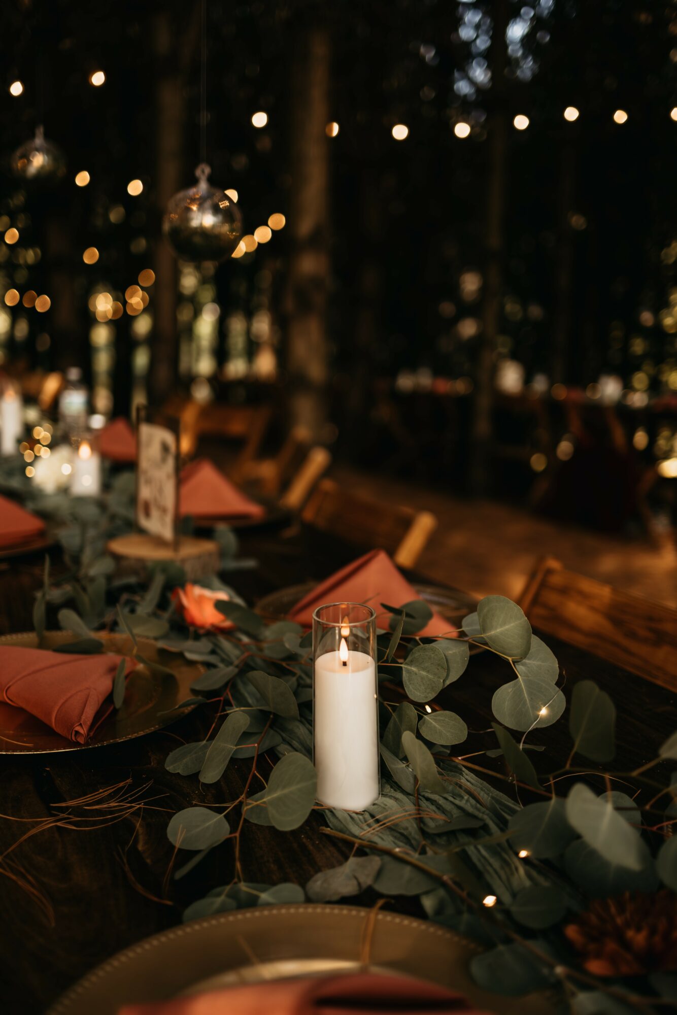 Table decorated with candles, greenery, and orange napkins outdoors at night with string lights overhead.