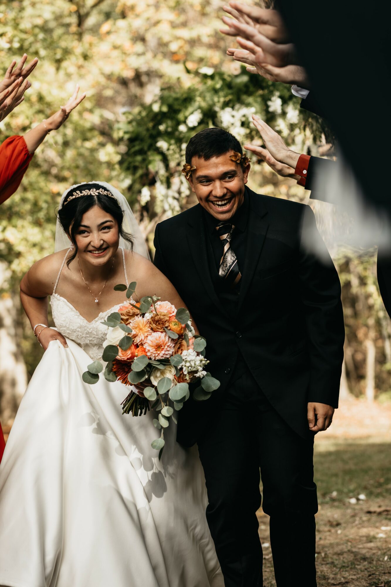 Smiling bride and groom outdoors, surrounded by people clapping, bride holding a bouquet, trees in background.