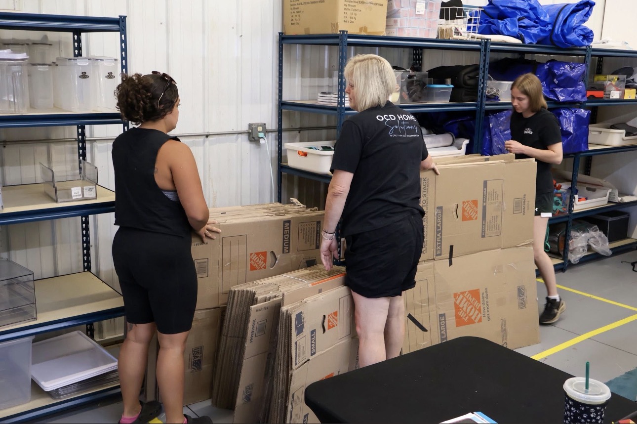 Three women work with cardboard boxes on shelves in a warehouse or storage room.