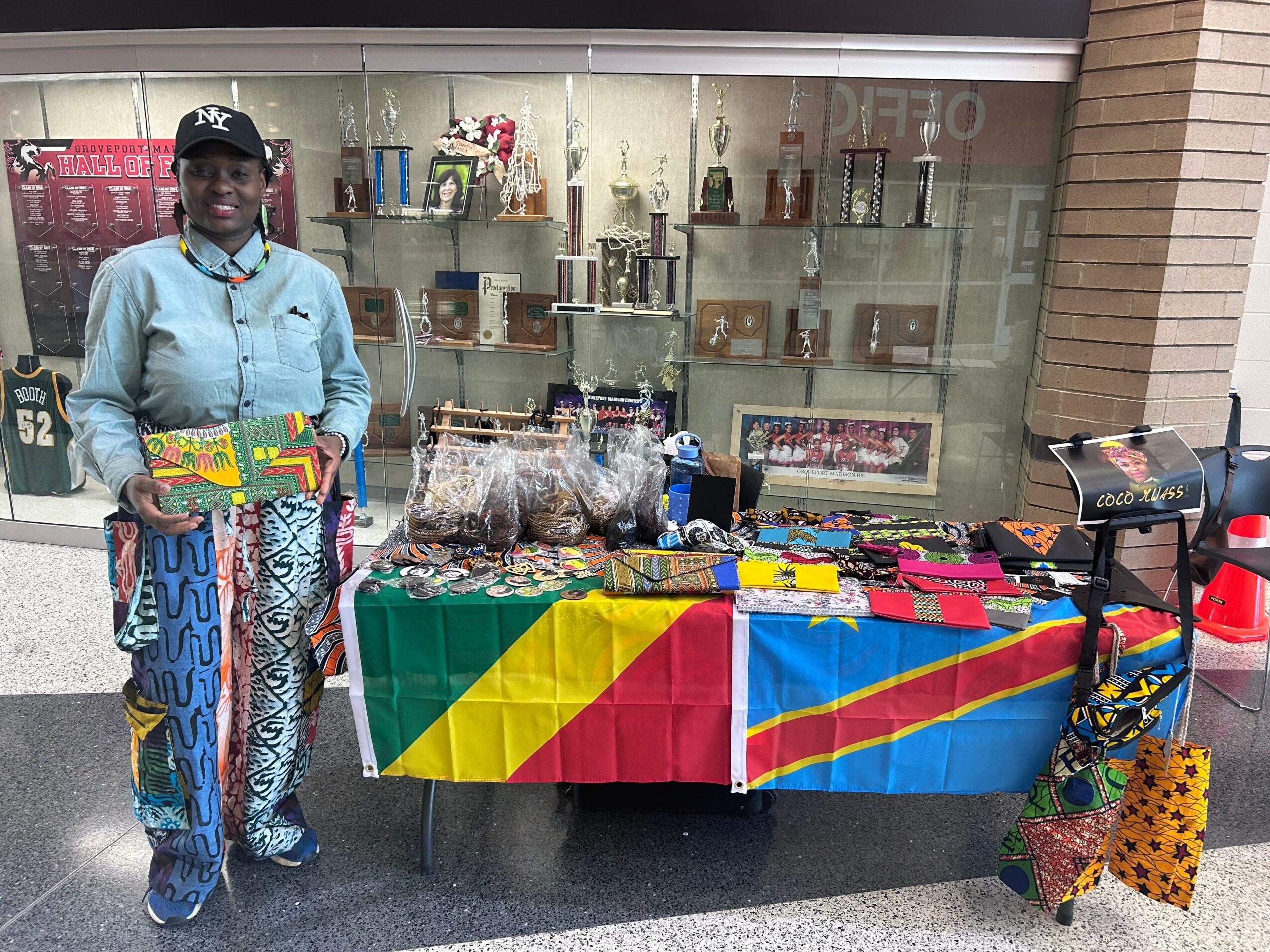 Person standing next to a table with colorful items, in front of a glass display case, holding a box, with a multicolored tablecloth.
