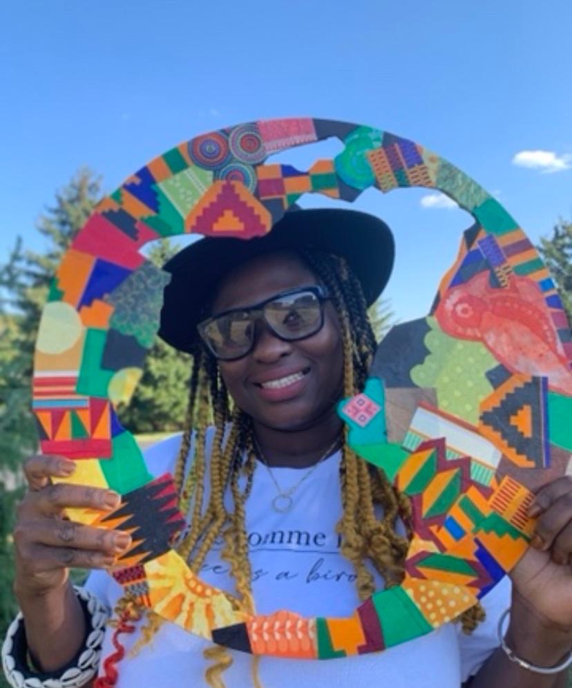 Person wearing glasses and a hat holding a colorful circular frame outdoors with trees and blue sky.