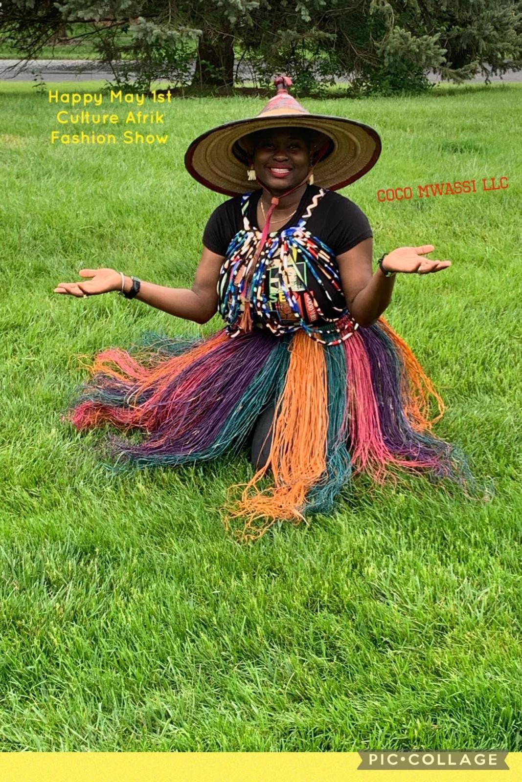 Person smiling outdoors wearing a large straw hat, colorful beaded necklace, and a multicolored skirt, with arms outstretched.