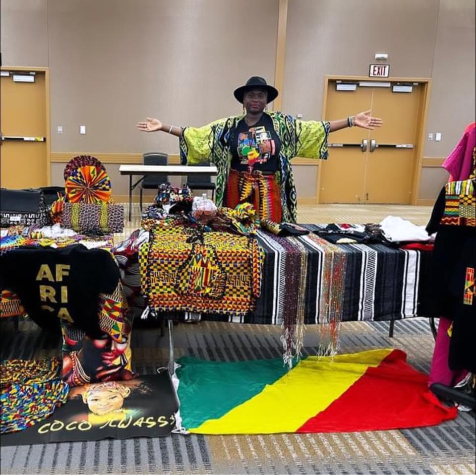 Woman standing behind a table with colorful African textiles and crafts, arms outstretched, in a room with yellow doors.