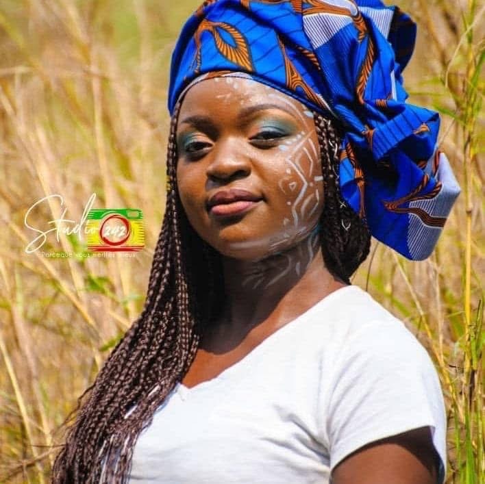 Woman with blue headwrap and face paint outdoors, wearing a white shirt, in a grassy field.