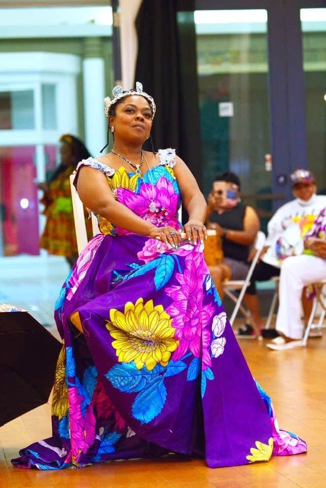 Woman in colorful dress with floral patterns, sitting with hands on lap, indoors with seated audience in background.