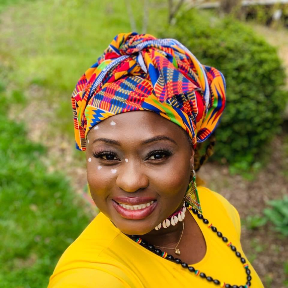 Woman smiling outdoors wearing a colorful headwrap and yellow top, with greenery in the background.
