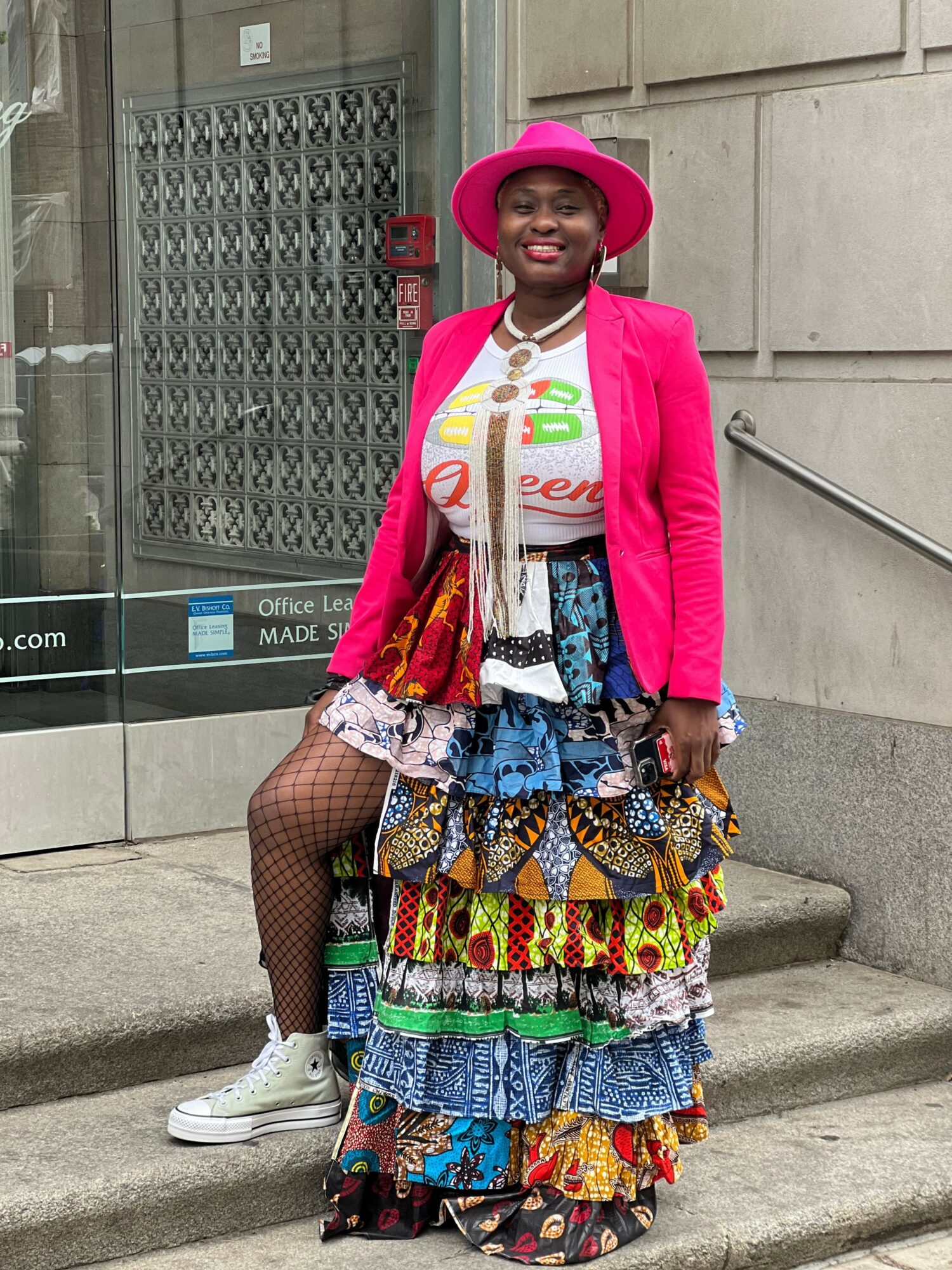 Person standing outdoors wearing a pink hat, pink jacket, and colorful patterned skirt, holding a bag, with stairs and building in background.