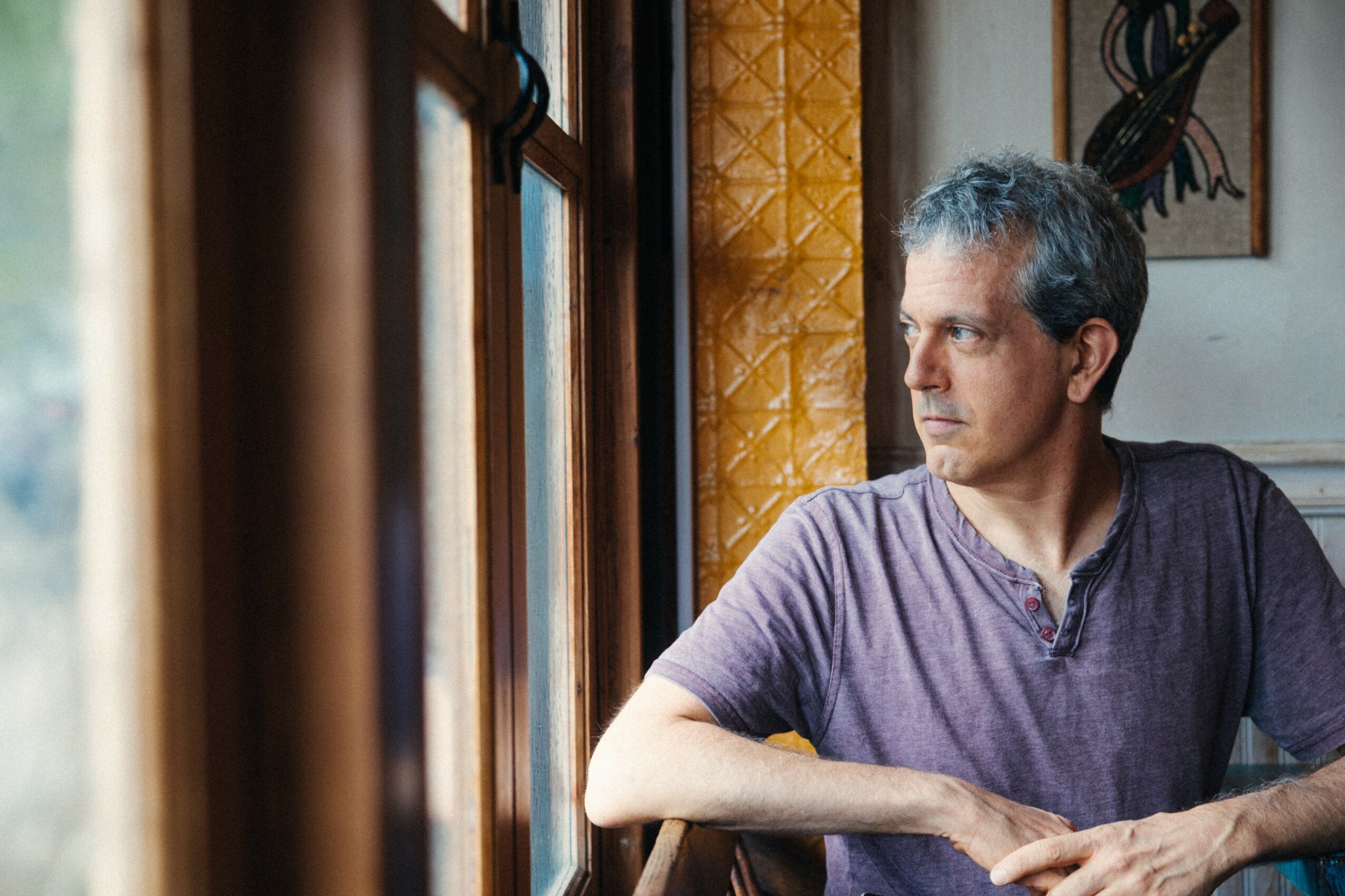 Man with gray hair wearing a purple shirt sitting by a window, looking outside, with curtains and wall art in background.