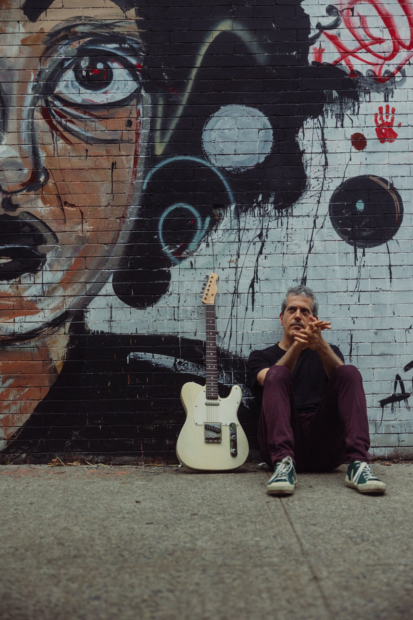 Man sitting on the ground with a guitar, graffiti wall background, one eye painted, holding a cigarette, wearing glasses and purple pants.