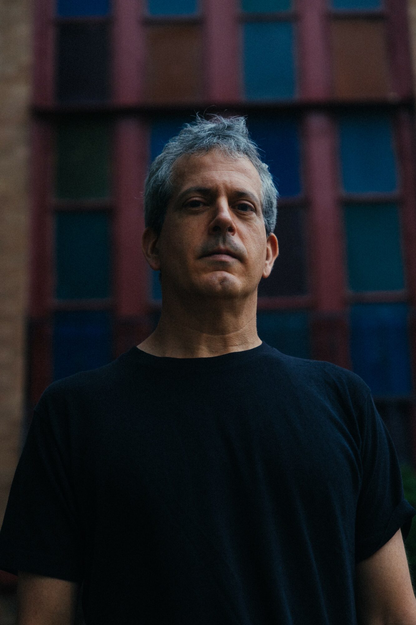 Man with gray hair and serious expression stands outdoors in front of a colorful building.