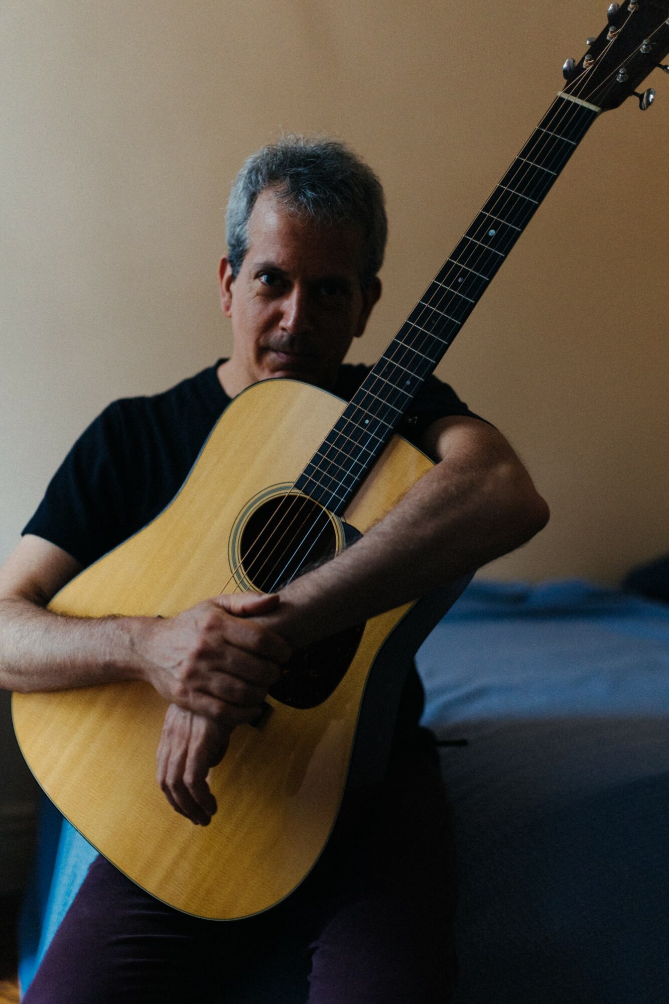 Person holding an acoustic guitar, sitting indoors with a neutral background, looking at the camera.