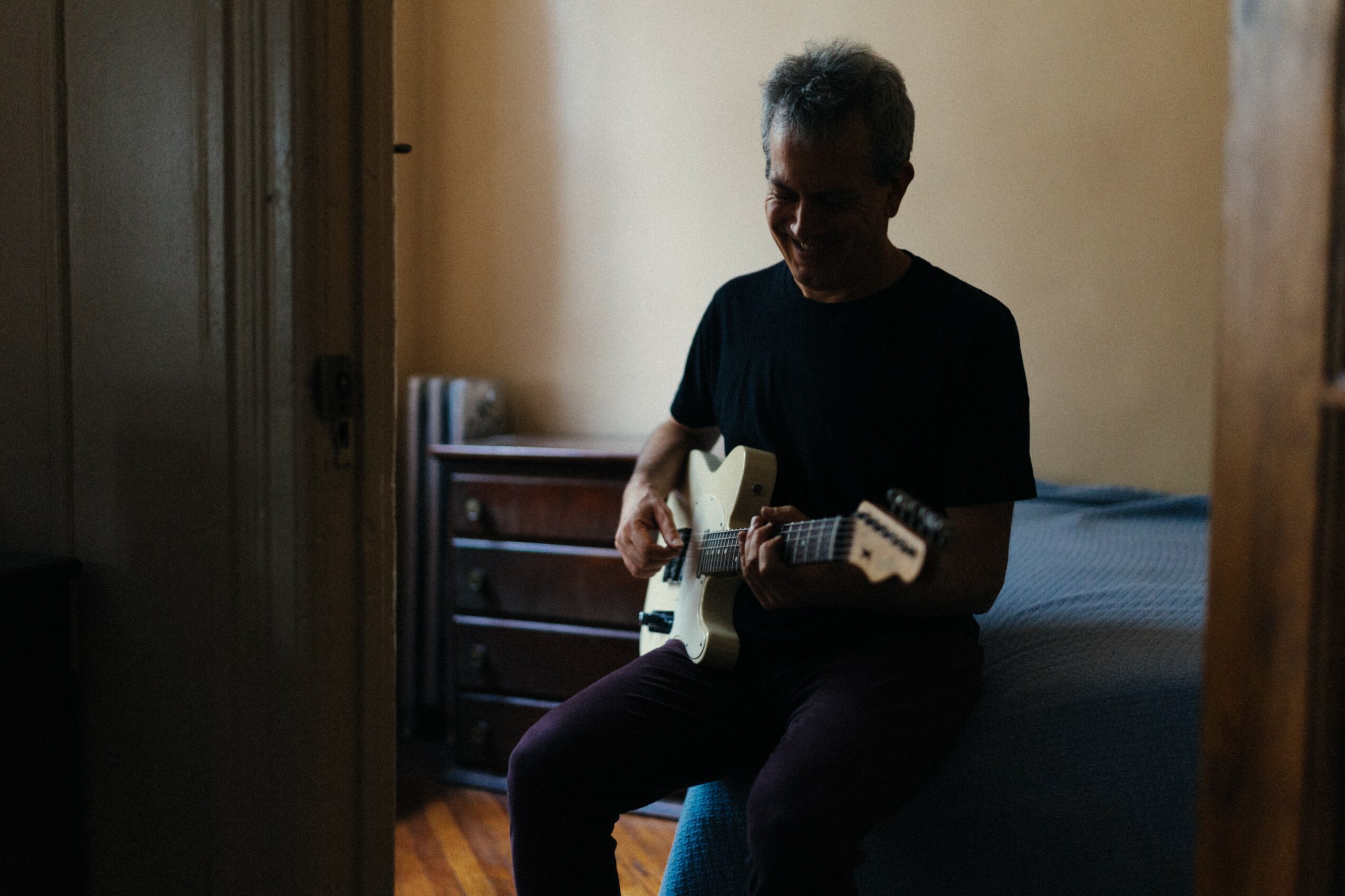 Person sitting on bed playing an electric guitar in a dimly lit room, with a dresser in the background.