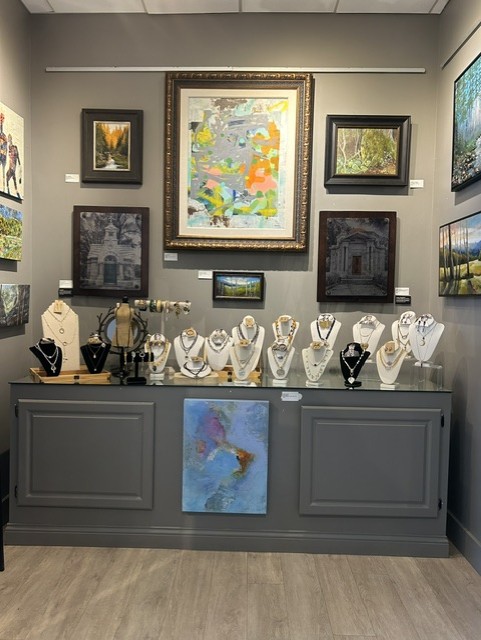 Gallery wall with framed paintings and a display of jewelry on bust stands above a gray cabinet, with a painting leaning against it.