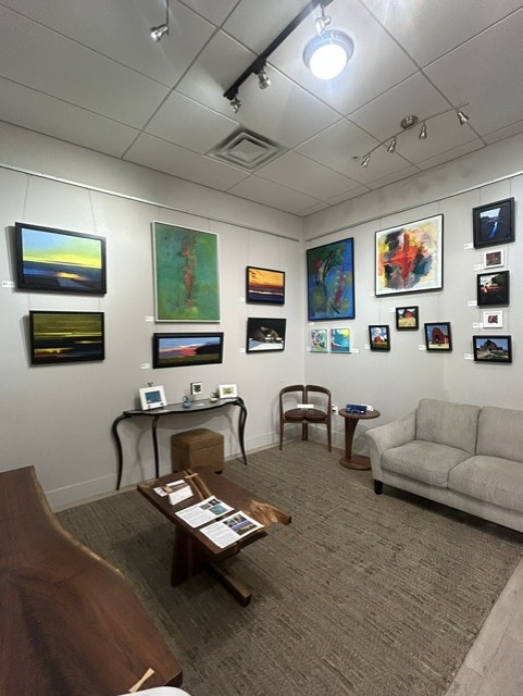 Gallery room with paintings on white walls, a sofa, a wooden table, and a chair, illuminated by ceiling lights.