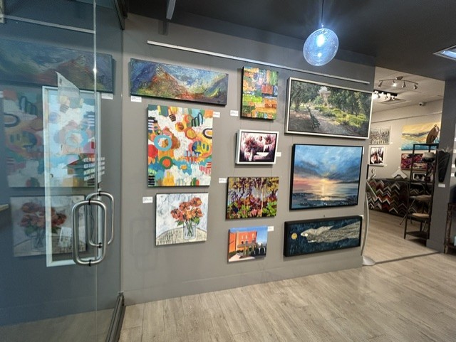 Gallery wall with various landscape and abstract paintings, glass door on the left, wooden floor, ceiling light fixture, and part of a store in the background.