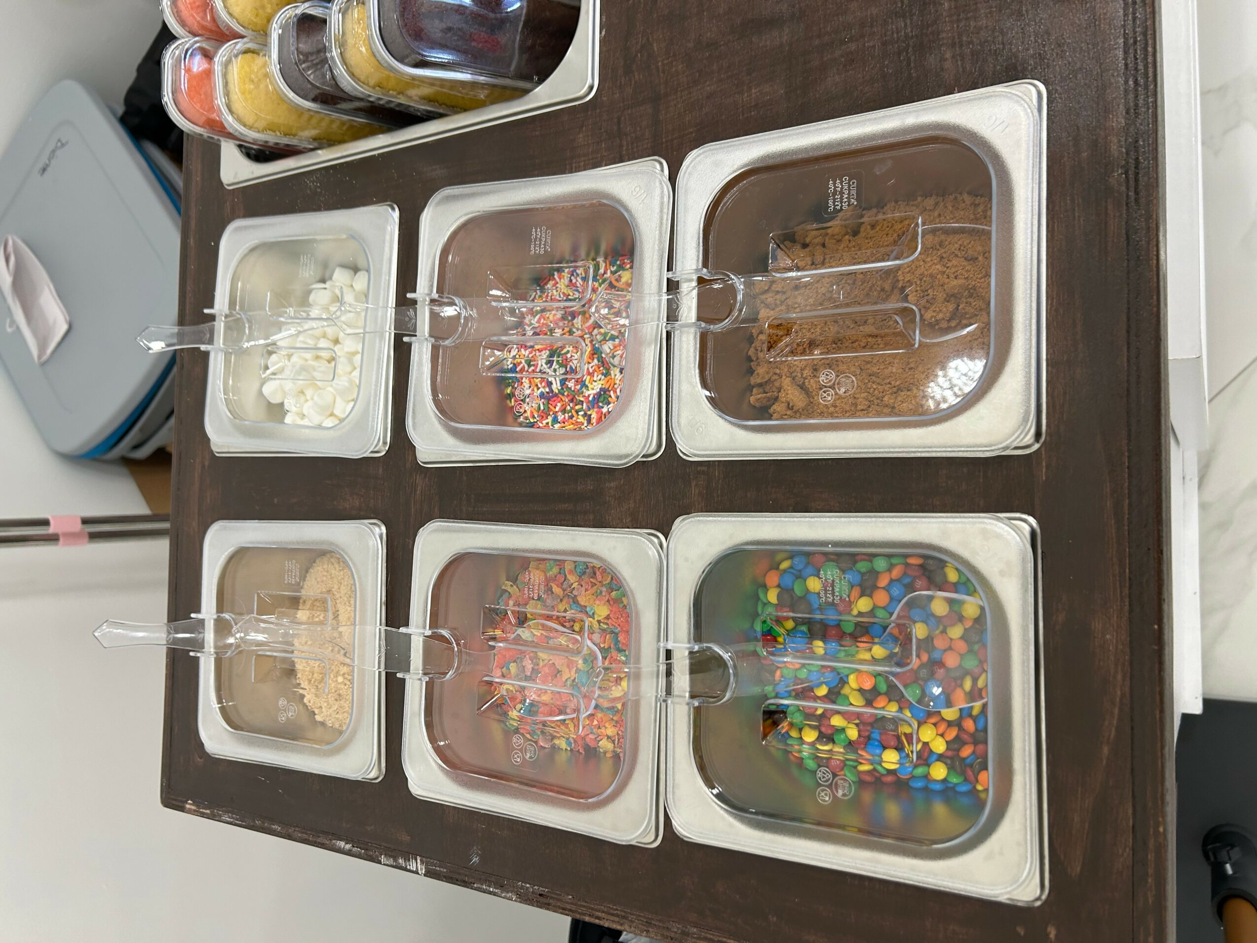 Six containers of decorated cookies and candies on a table, with some containers containing colorful sprinkles and toppings.