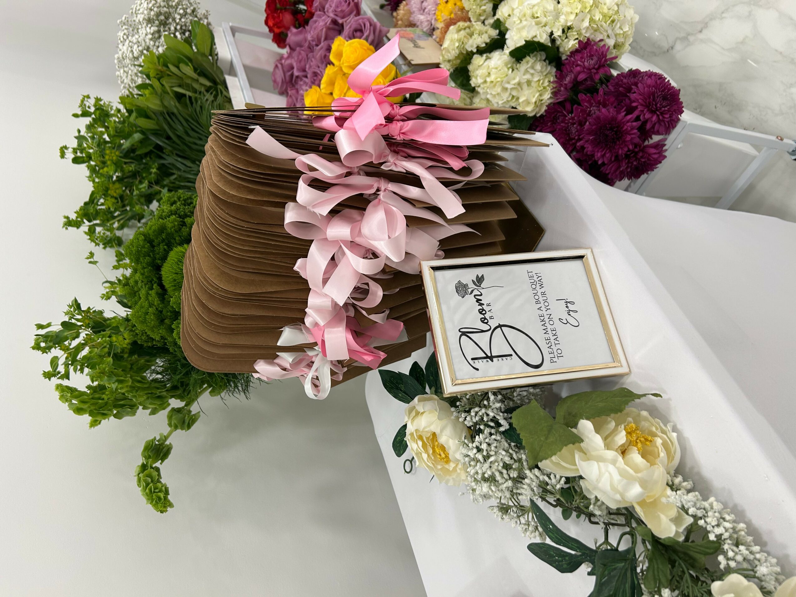 A large chocolate cake decorated with pink ribbons, surrounded by colorful flowers and a small floral arrangement on a white table.
