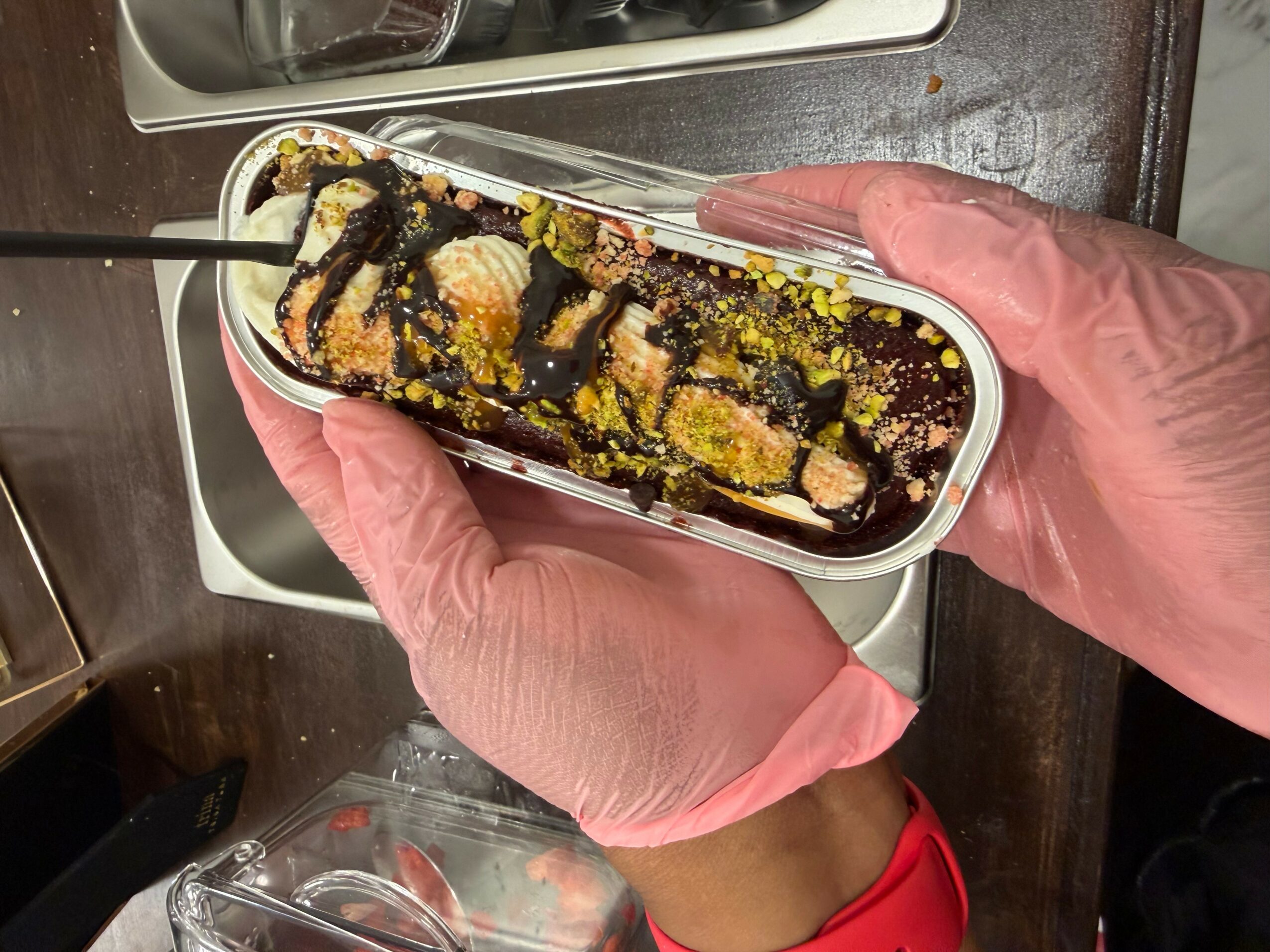 Person holding a container of sushi with chocolate drizzle and yellow topping, wearing pink gloves, over a stove.