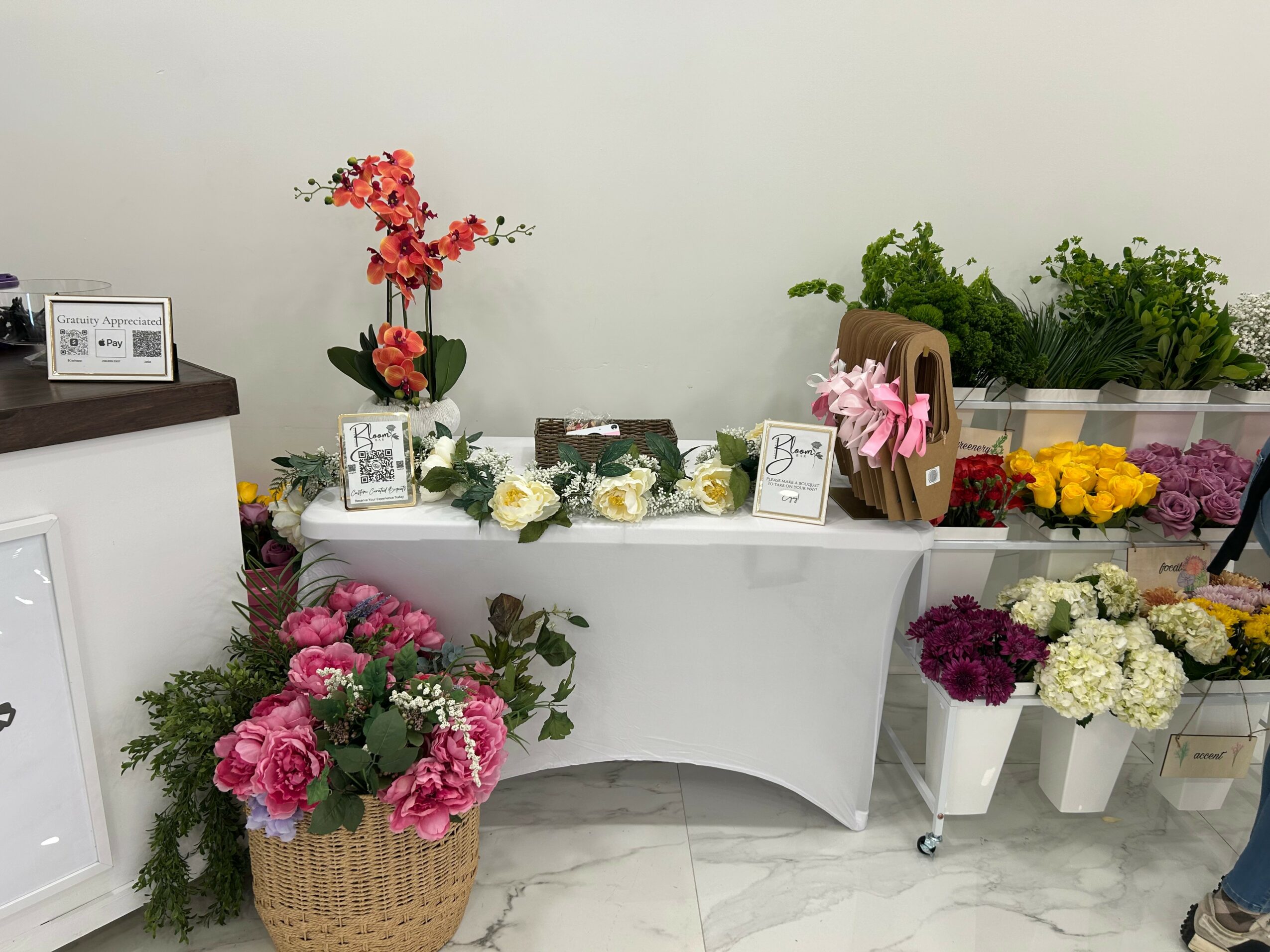 Display of various flowers and plants on tables and shelves, with framed photos and signs, against a plain wall.