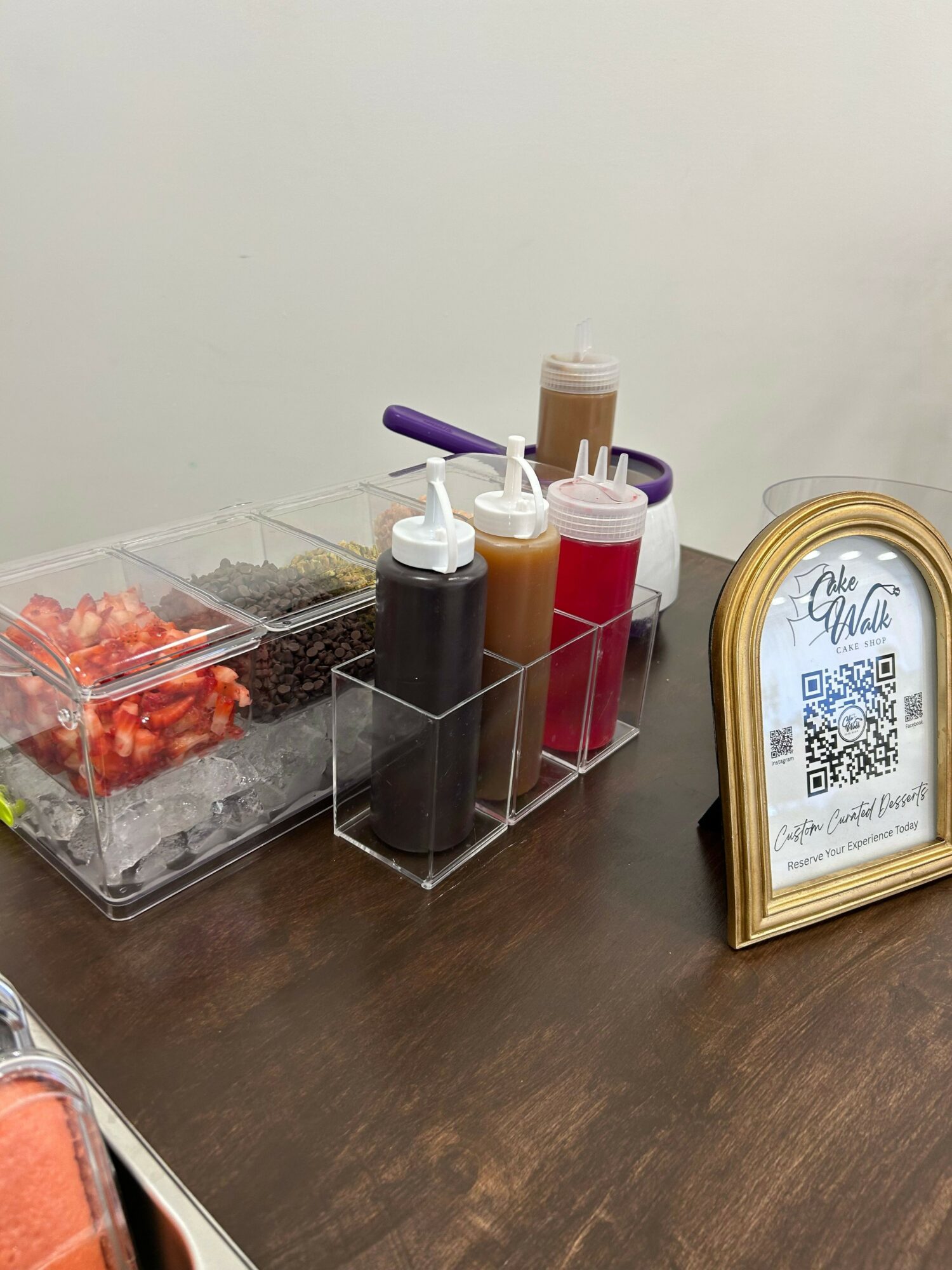 Clear container with chopped strawberries, bottles of sauces, and a purple spoon on a wooden surface, with a framed QR code in the background.
