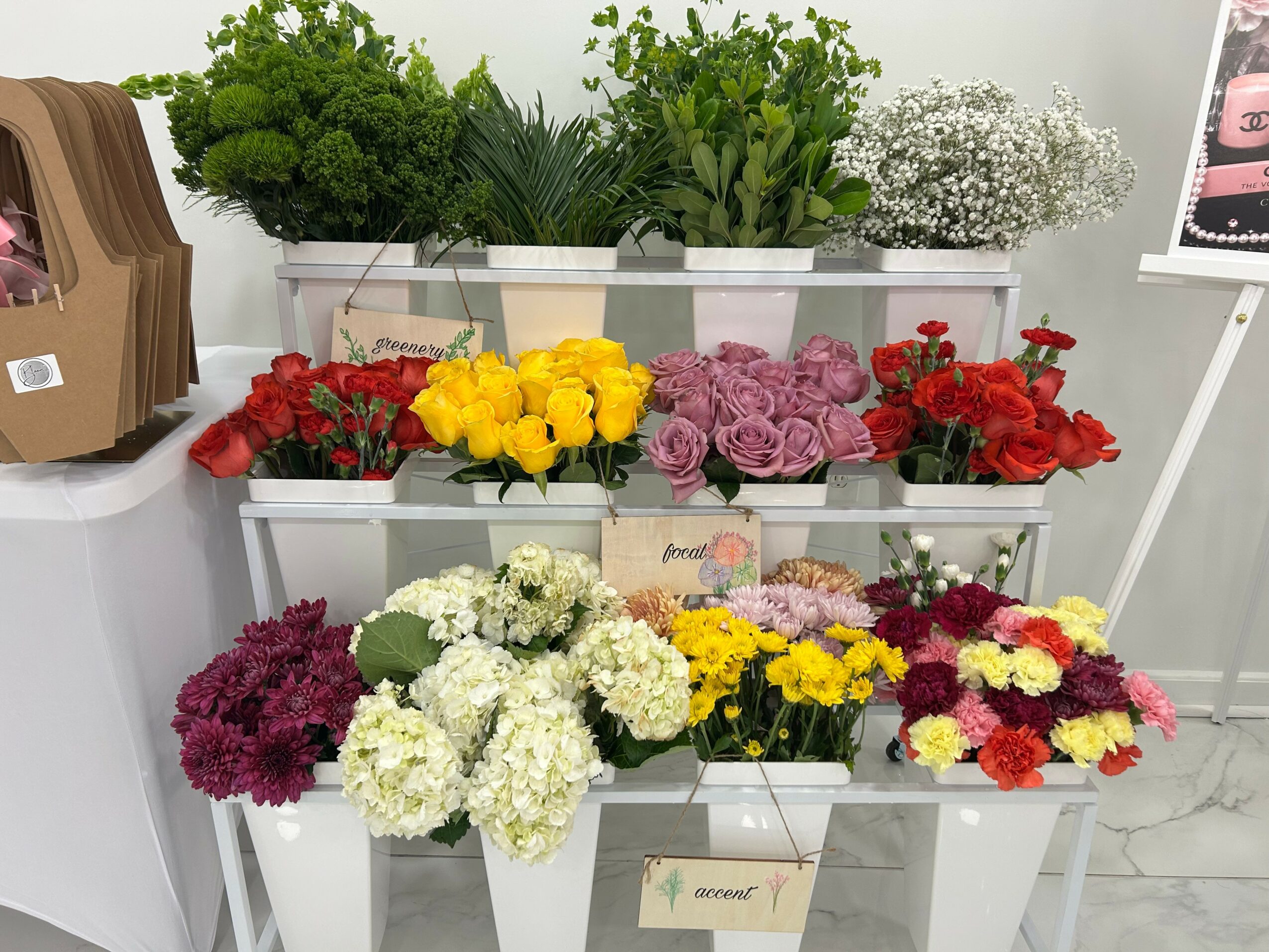 Display of colorful flowers including red, yellow, purple, white, and pink arranged on white shelves with green plants in background.