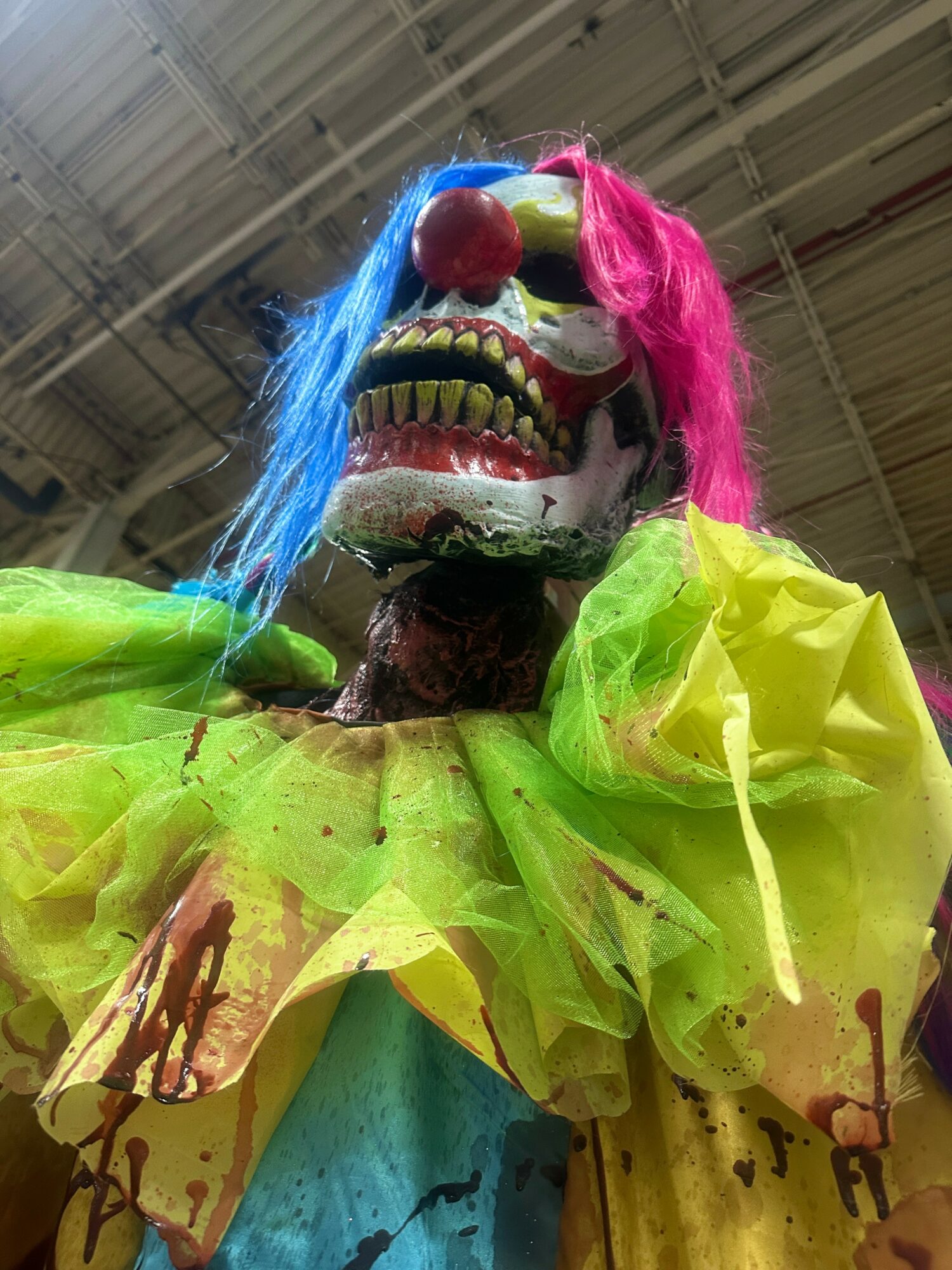 Colorful clown-like figure with a skull face, pink and blue hair, and a bright yellow and green costume, hanging from ceiling.