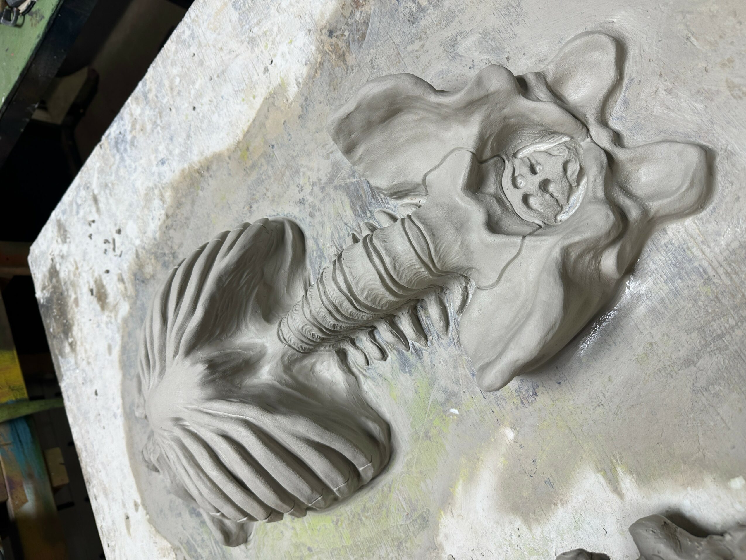 Clay sculpture of a mermaid with a fish tail, lying on her side with arms behind her head, on a concrete surface.