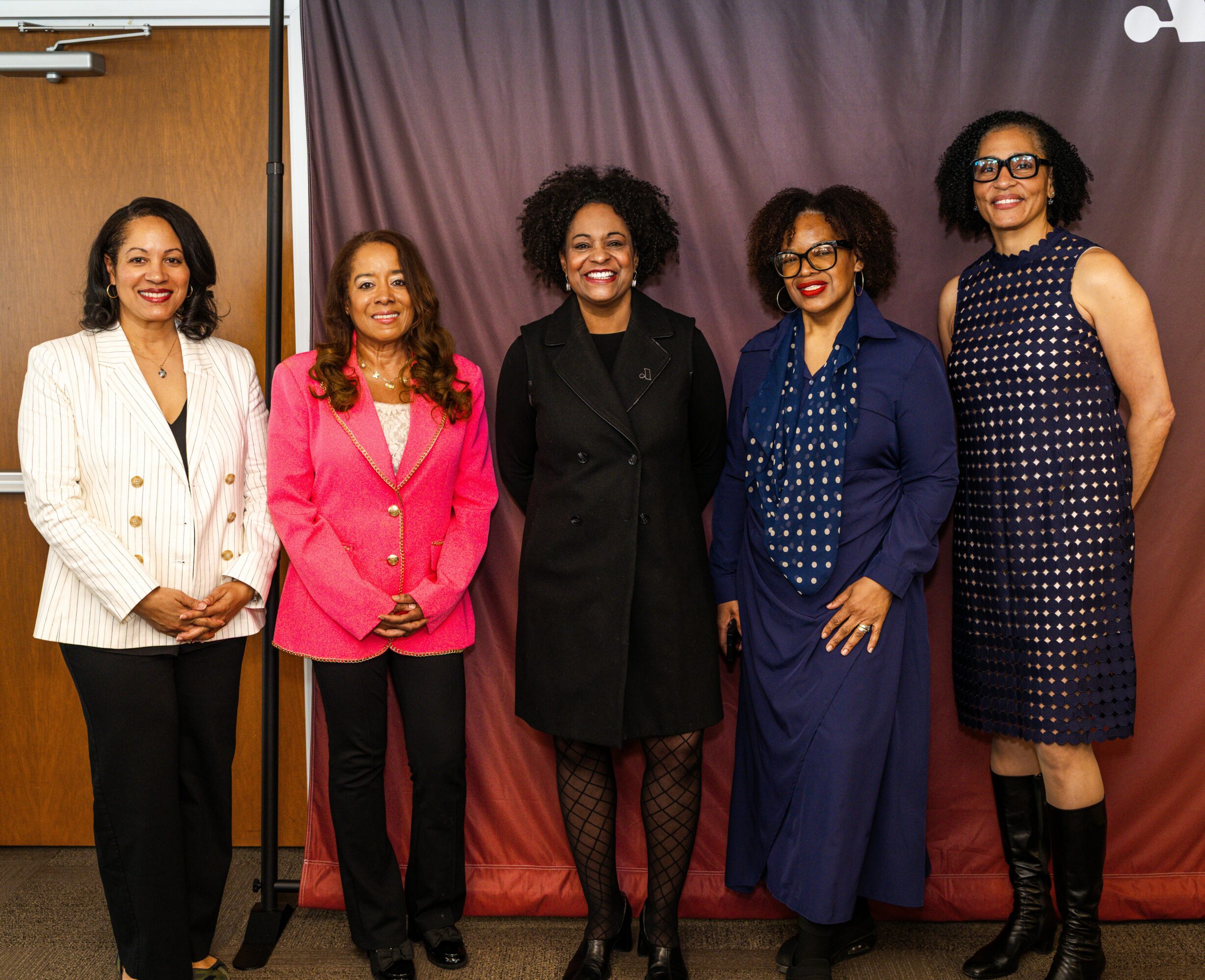 Five women standing together in front of a purple curtain, smiling, dressed in formal attire.
