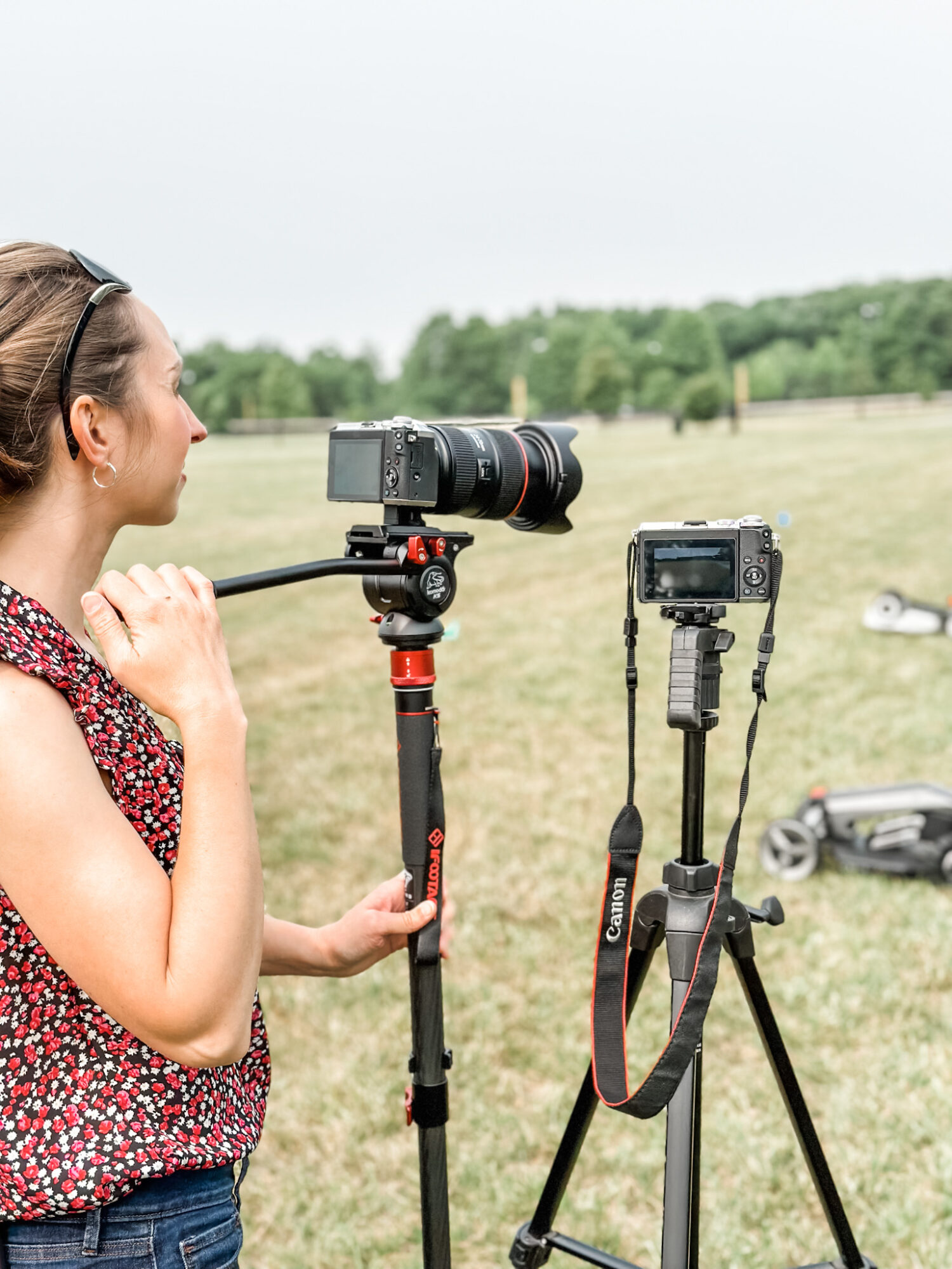Woman with camera on tripod outdoors in a grassy field, with another camera on a tripod nearby, overcast sky.