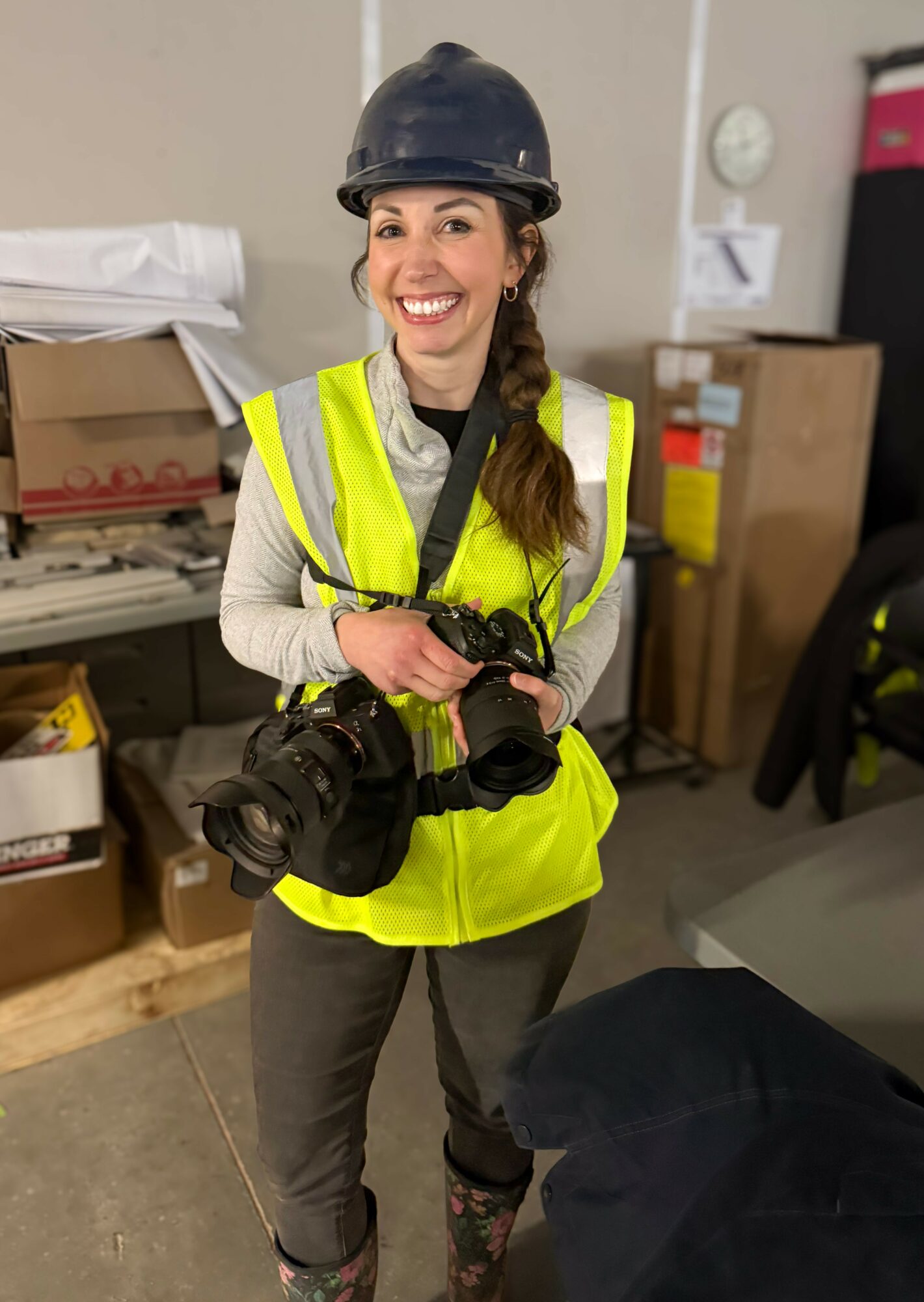 Smiling woman in safety vest and helmet holding two cameras indoors, with storage boxes and equipment in background.