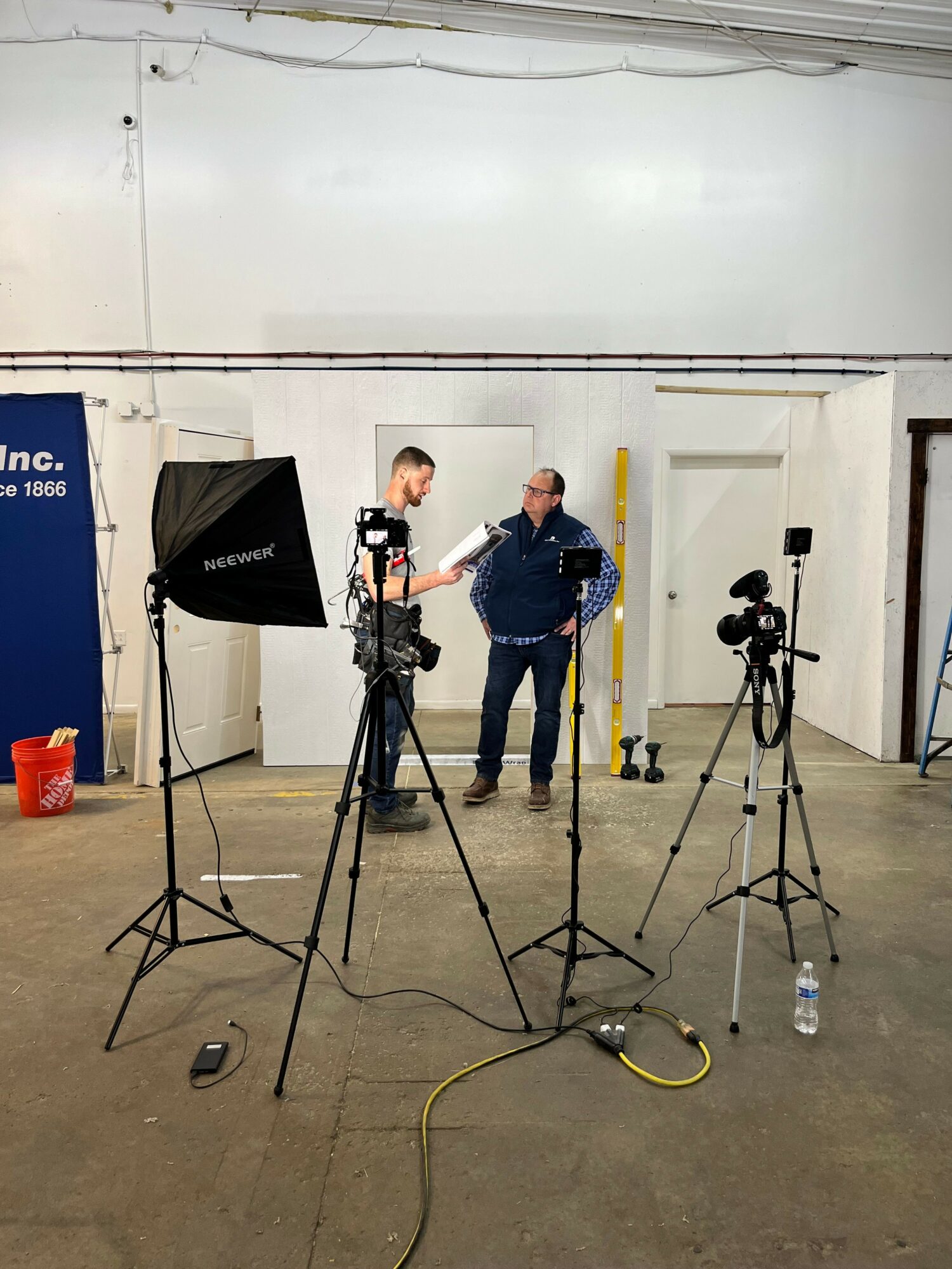 Two people in a studio with camera equipment, standing near a large light and a white door, concrete floor.