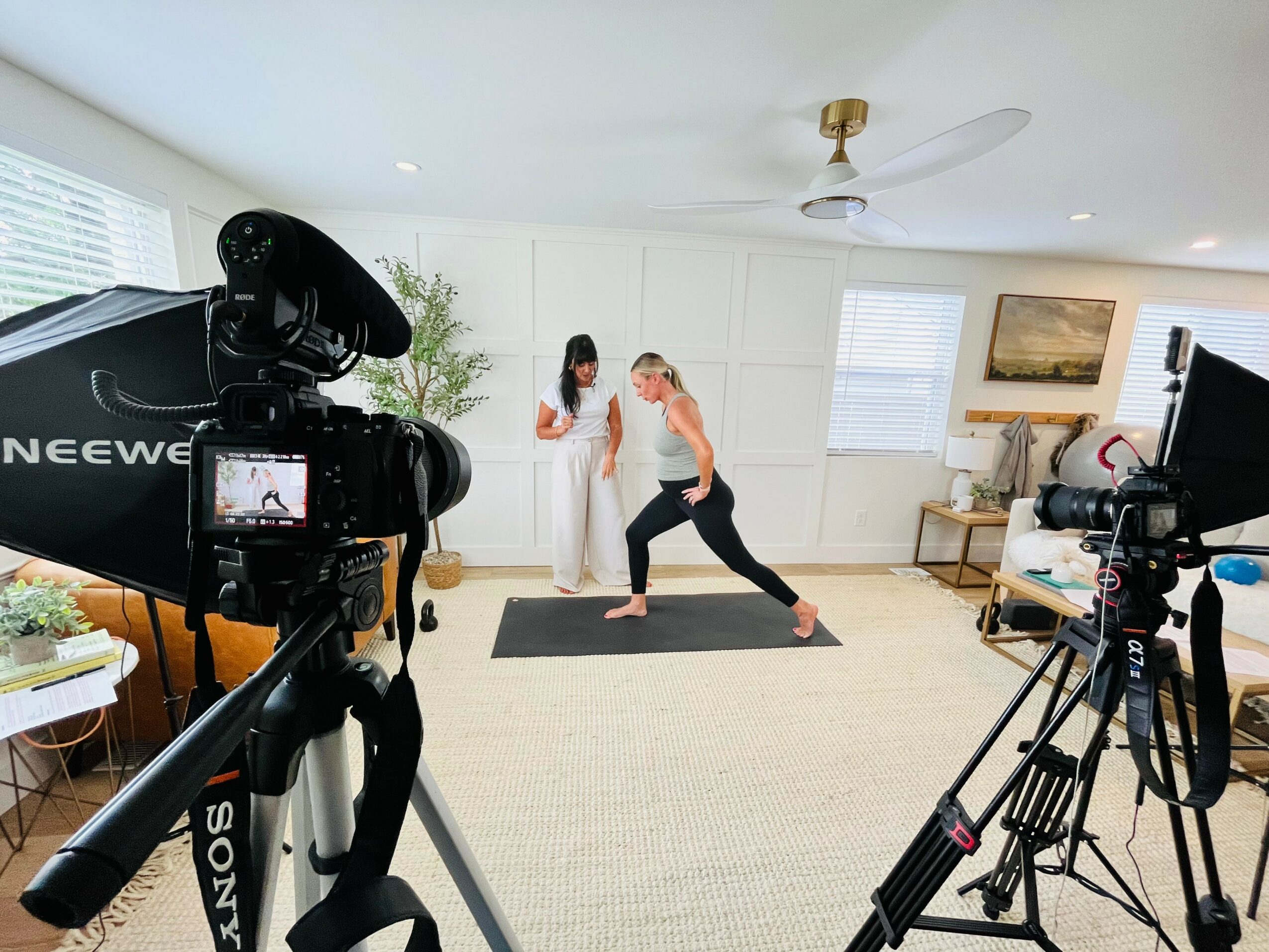 Two women practicing yoga in a bright living room, with cameras recording the session, one woman in a white outfit and the other in black leggings and a tank top, on a black mat.