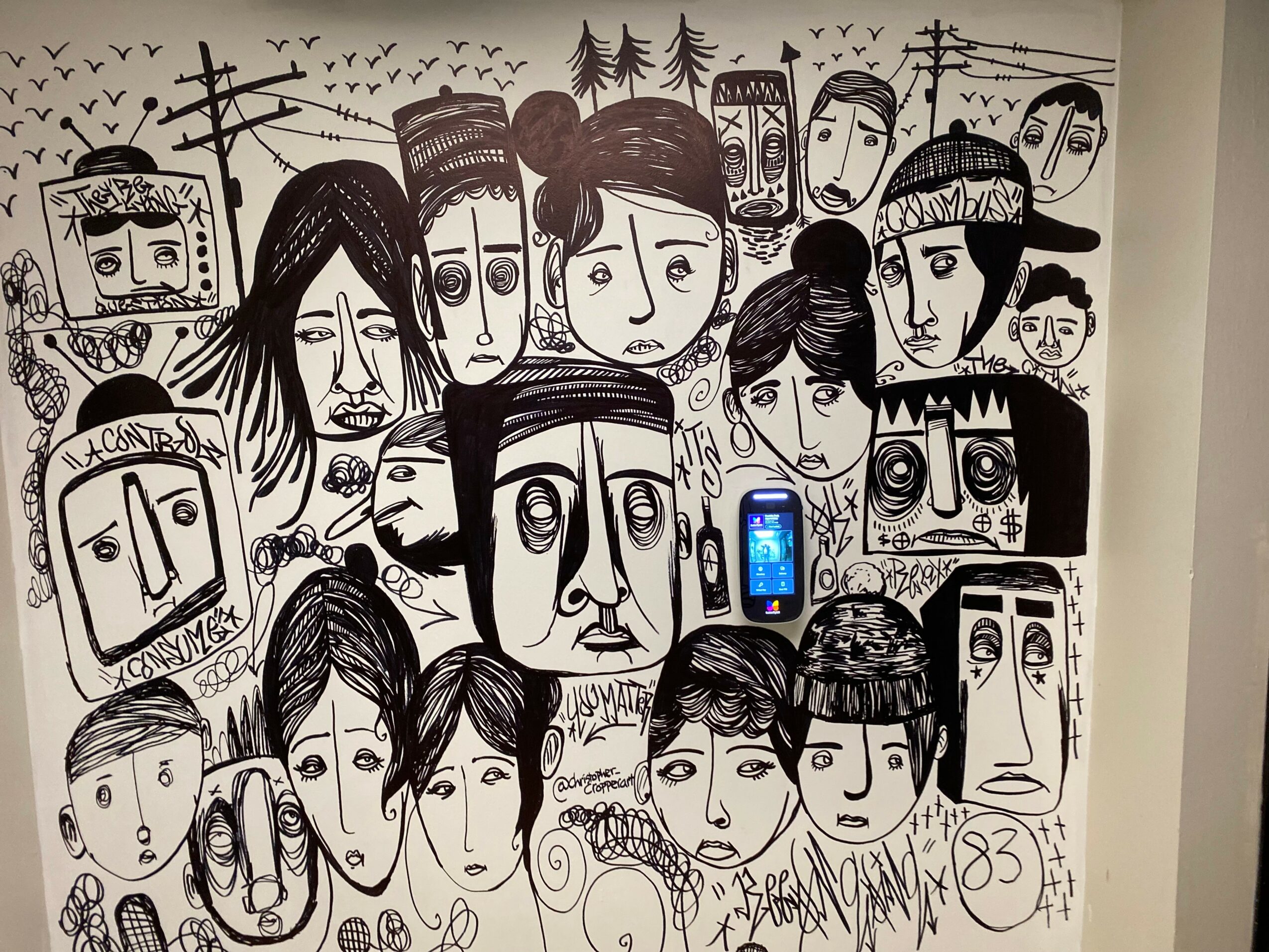 Wall mural with black and white cartoon faces of various expressions and hairstyles, some with phones, background with birds and trees.