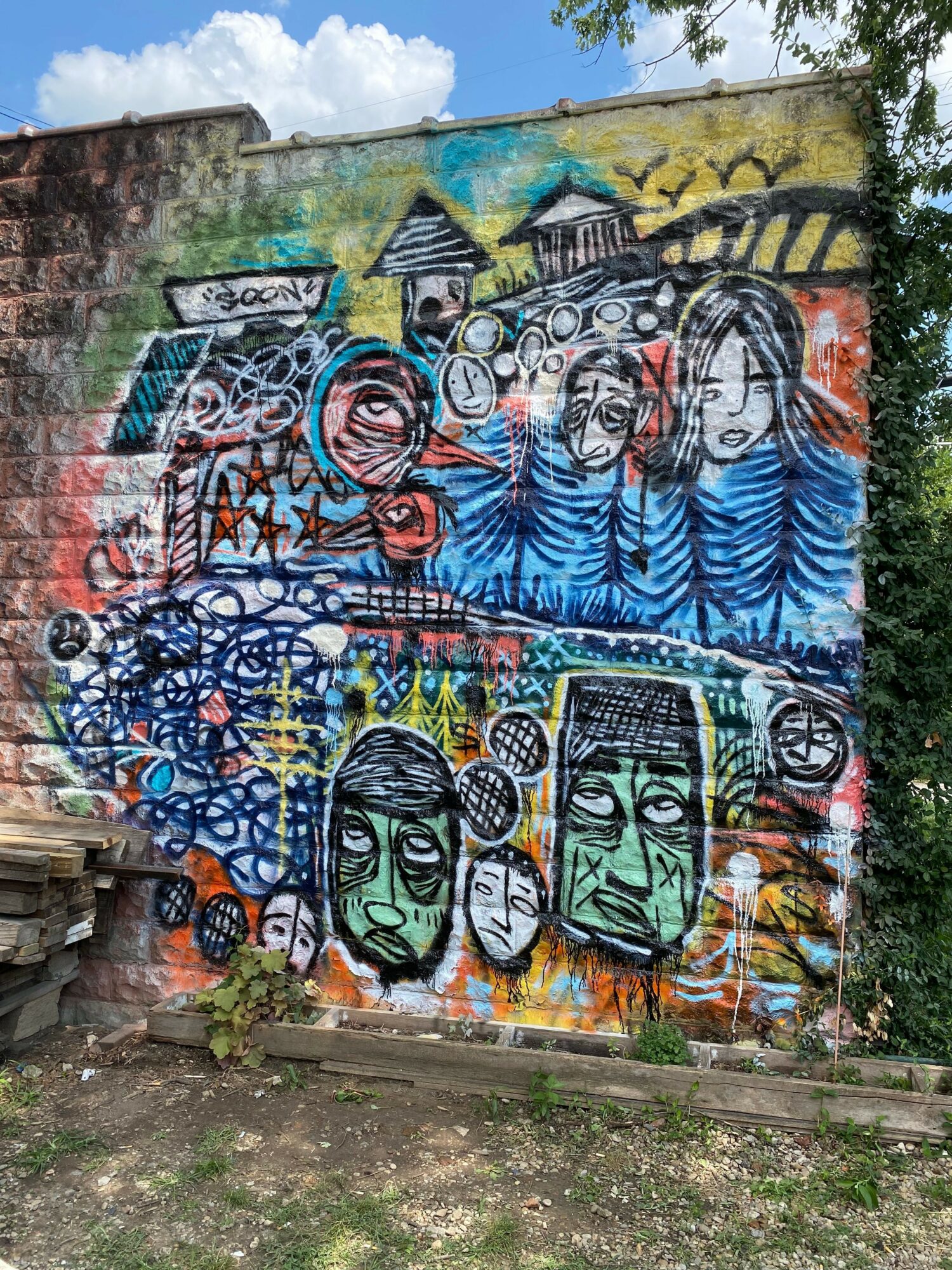 Colorful graffiti mural with abstract faces, shapes, and patterns on a wall, with trees and sky in the background.