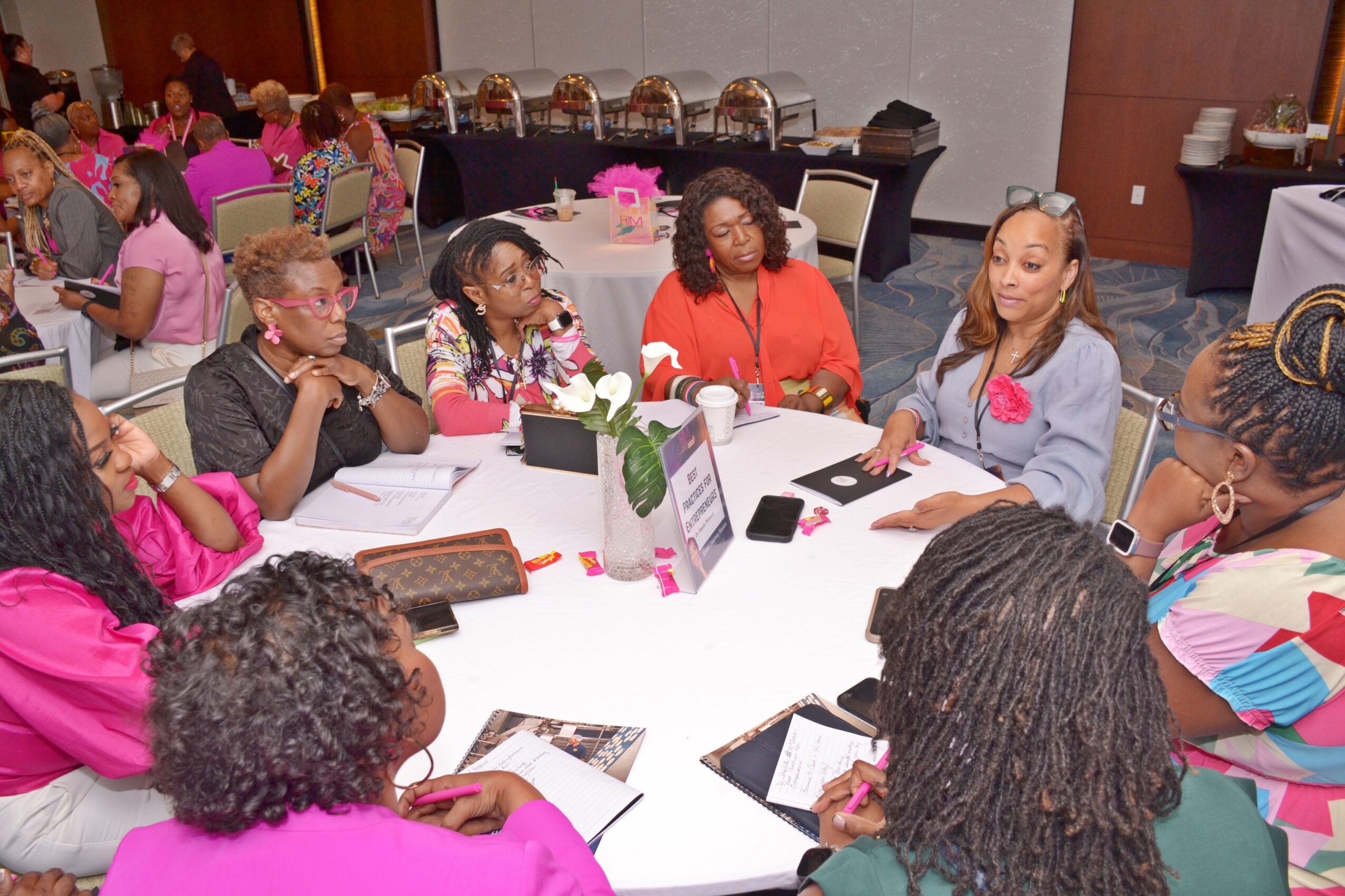 Group of women and girls sitting around a table with notebooks and tablets, in a large room with other groups in the background.