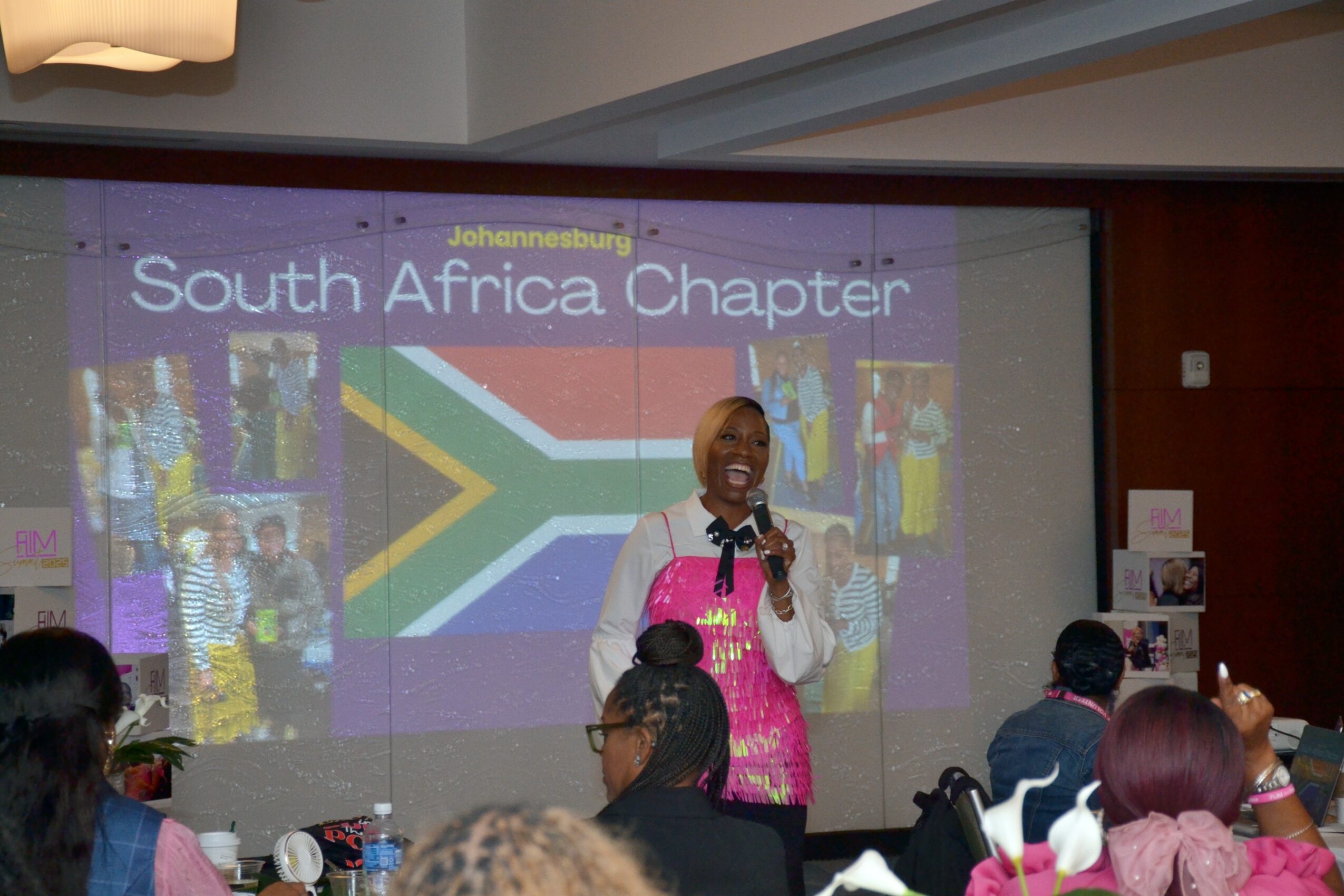 Woman speaking at South Africa chapter event with a large screen displaying the South African flag and images in the background.