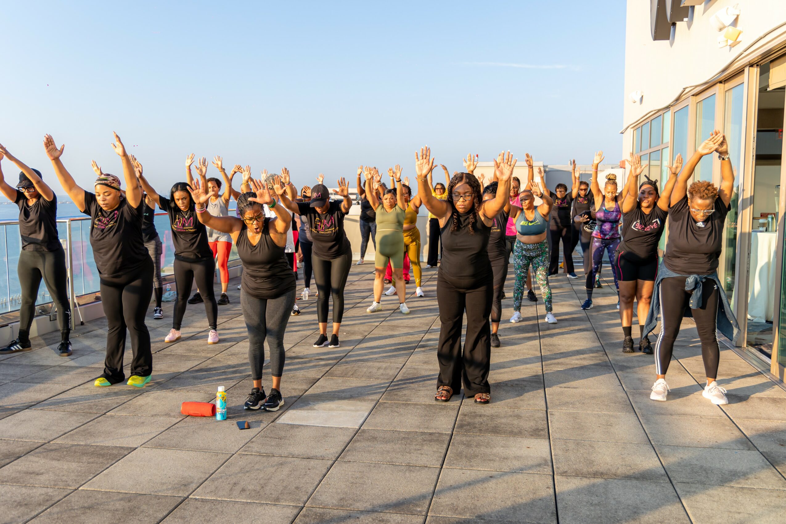 Group of people participating in outdoor exercise on a tiled terrace, raising arms, with a building and sky in background.