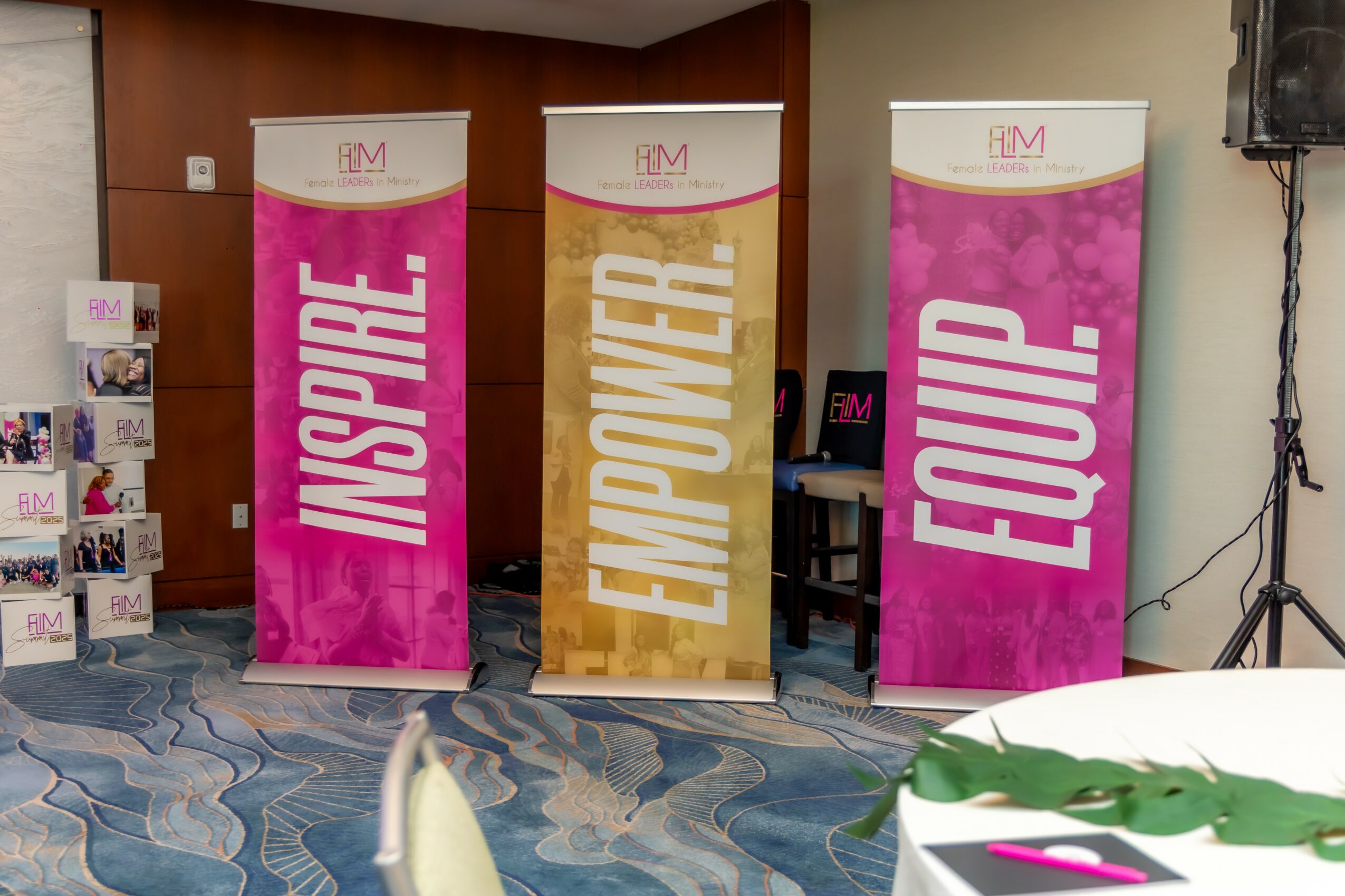 Three vertical banners with pink, gold, and pink backgrounds displaying the words 'INSPIRE,' 'EMPOWER,' and 'EQUIP' in large white letters, set indoors.