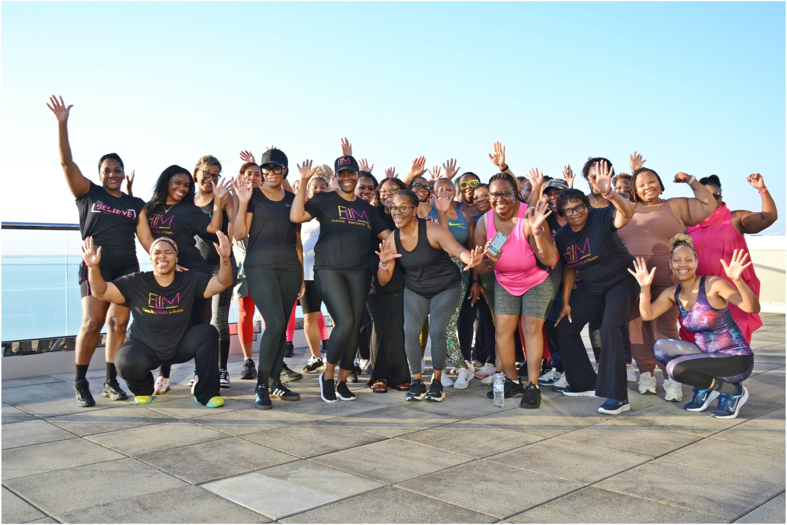 Group of people outdoors waving and smiling, standing on a paved surface with a clear blue sky.